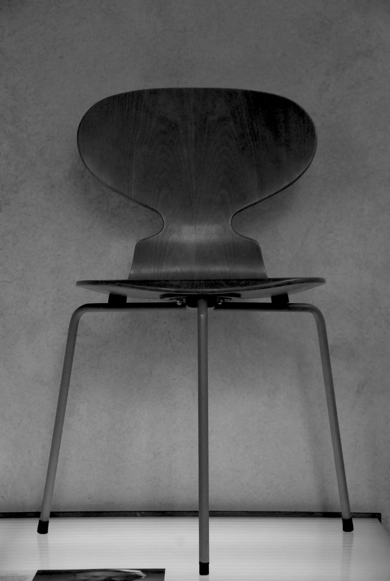 A frontal view of a dark plywood Ant Chair with a pinched waist and three slender steel legs against a plain wall.