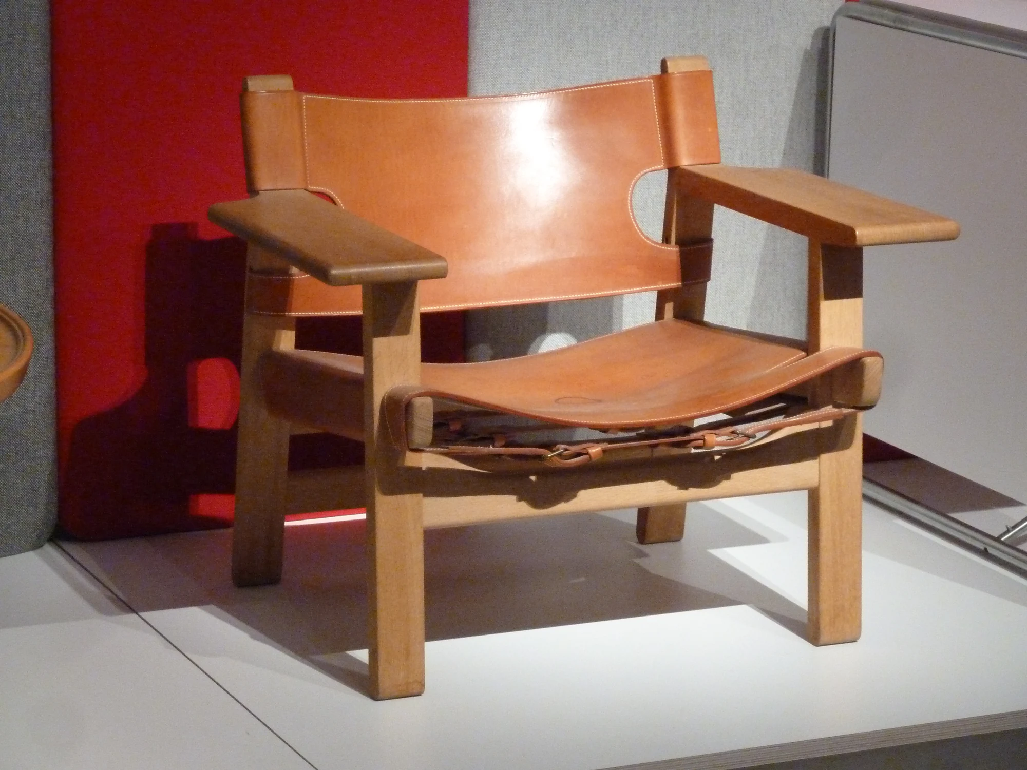 Three-quarter view of a low Spanish Chair with broad pale timber arms, a tan leather back and seat, and buckled leather straps visible beneath the front edge.