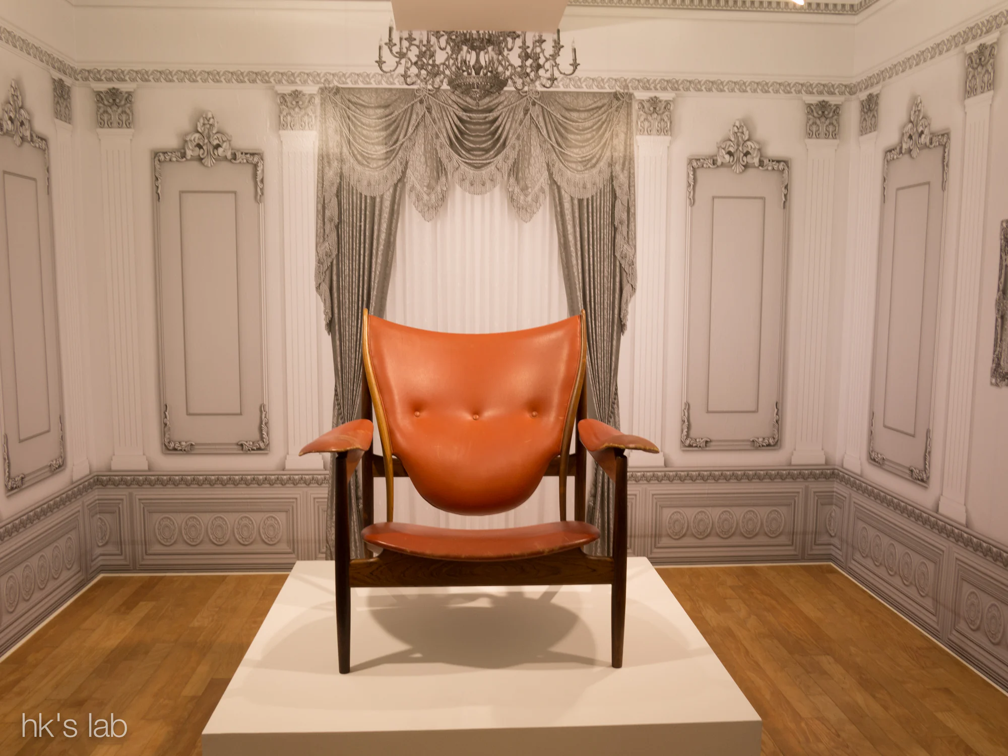 A frontal view of a Chieftain Chair with a broad orange leather back and seat suspended inside a dark timber frame in a later exhibition setting.