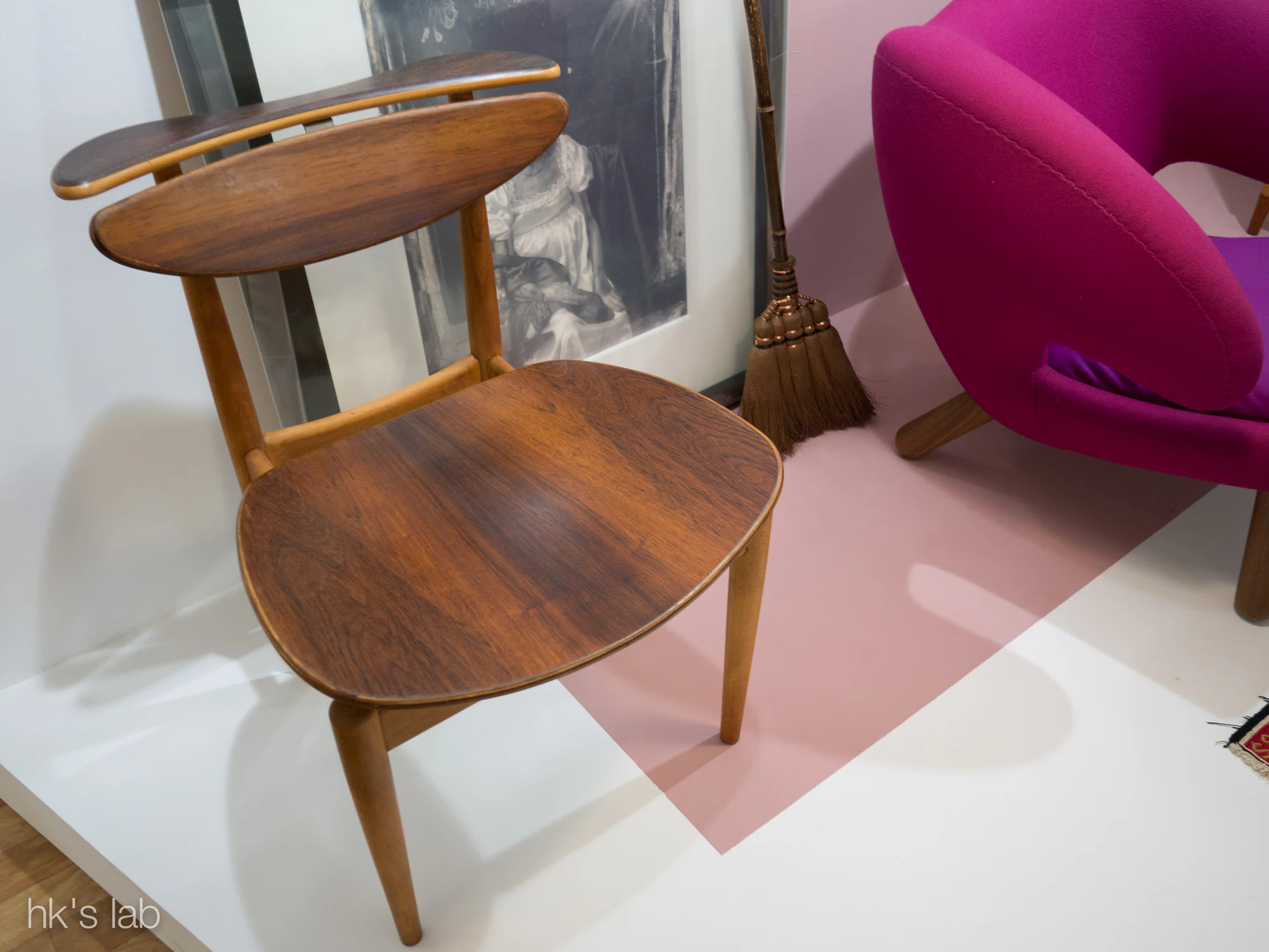 Angled exhibition view of a wooden Finn Juhl Reading Chair with a broad curved back board, a separate horizontal top rail and a rounded veneered seat.