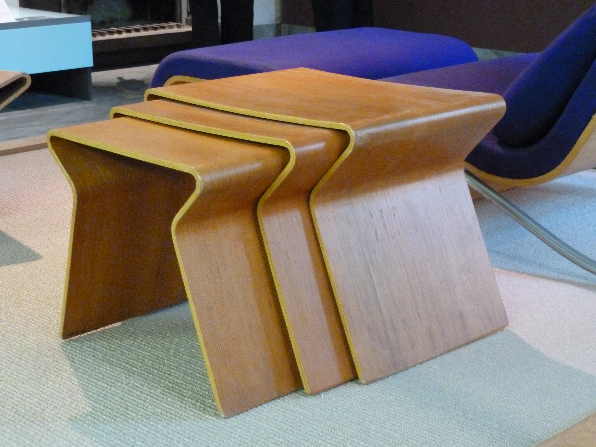 Three Grete Jalk nesting tables in graduated sizes, each made as a broad timber-coloured sheet that curves down at both ends, displayed with other furniture in a museum.