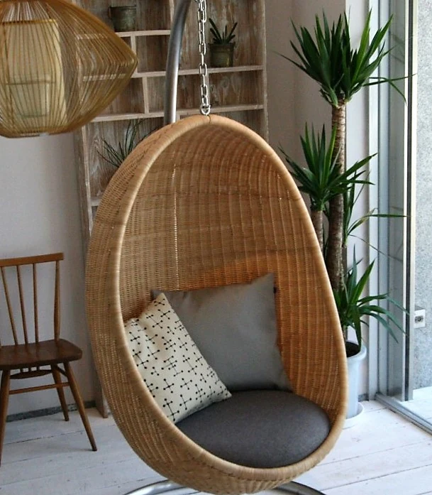 A natural rattan Hanging Egg Chair suspended by a chain from a curved stand in a bright domestic interior.