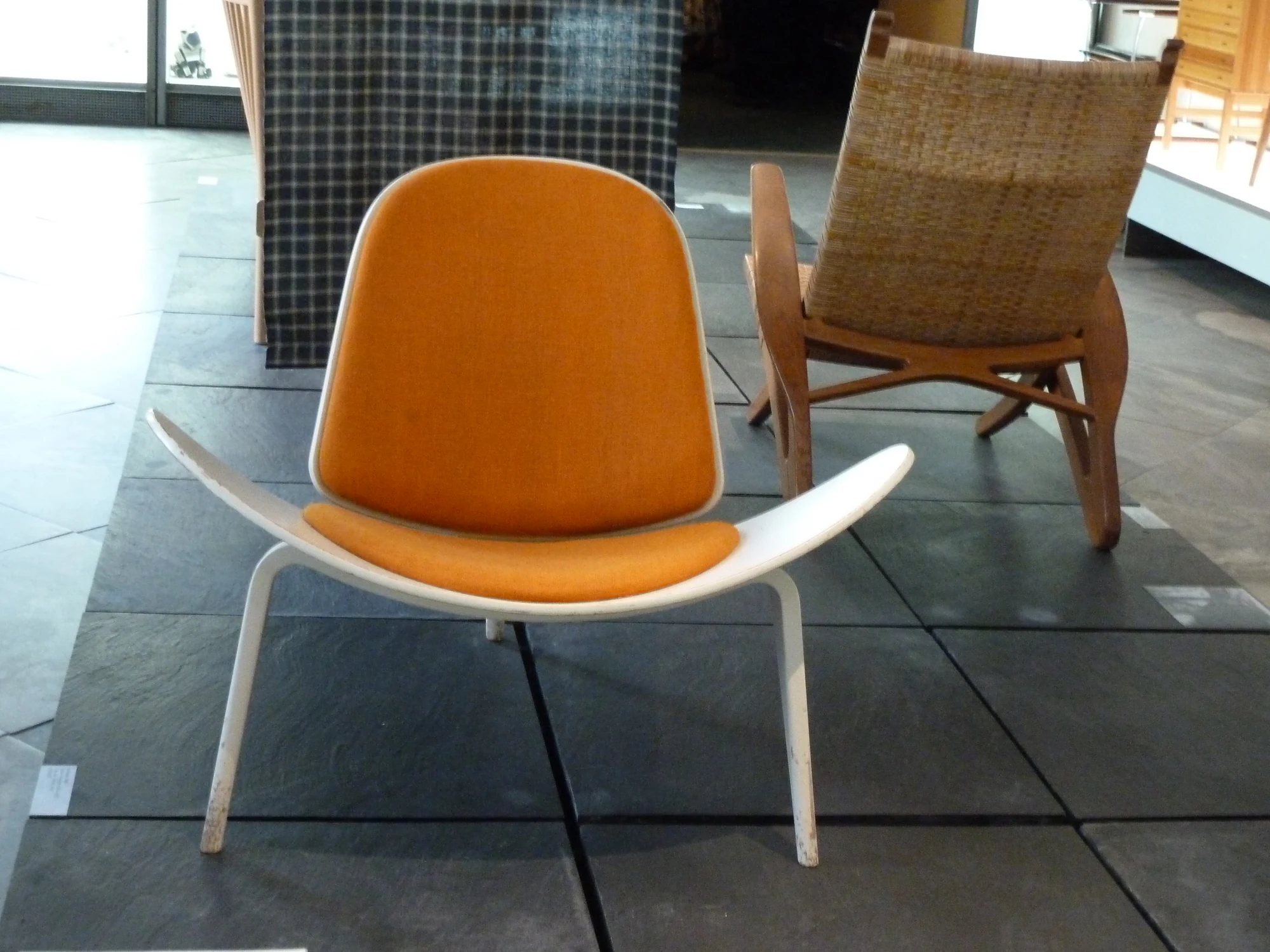 Front view of a Shell Chair with a wide white wing-shaped seat, a separate orange back pad and three pale legs, displayed beside other chairs in a museum gallery.