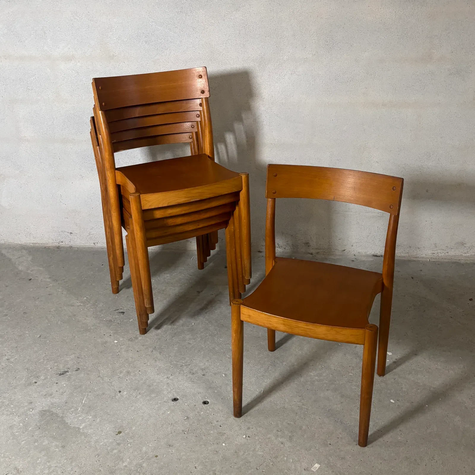 One Portex side chair stands in front of several matching chairs nested into a compact stack, all in warm-toned wood against a plain wall.