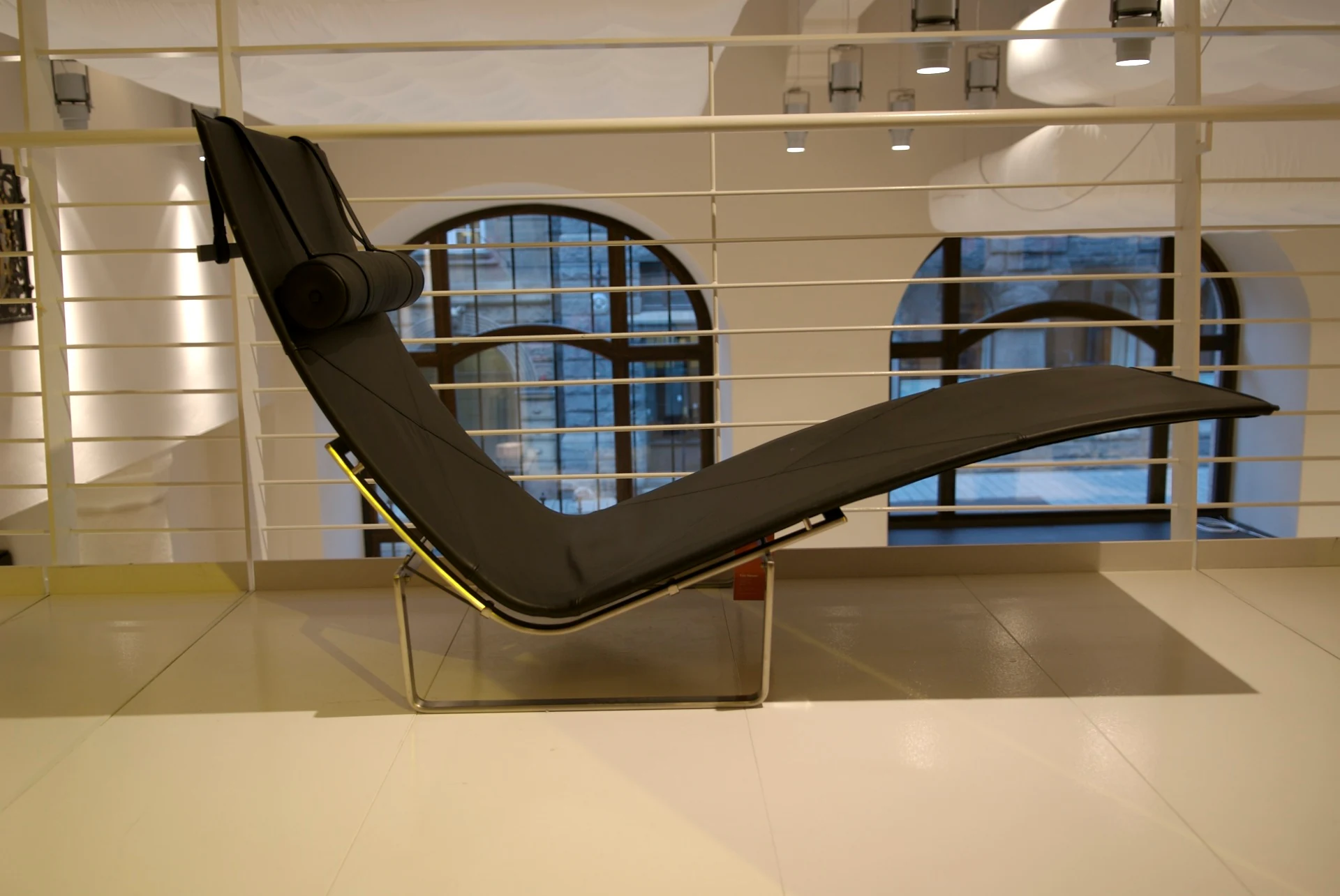 Side view of a black leather PK24 chaise longue, its long curved seat resting above a low rectangular stainless-steel base with a cylindrical headrest at the raised end.