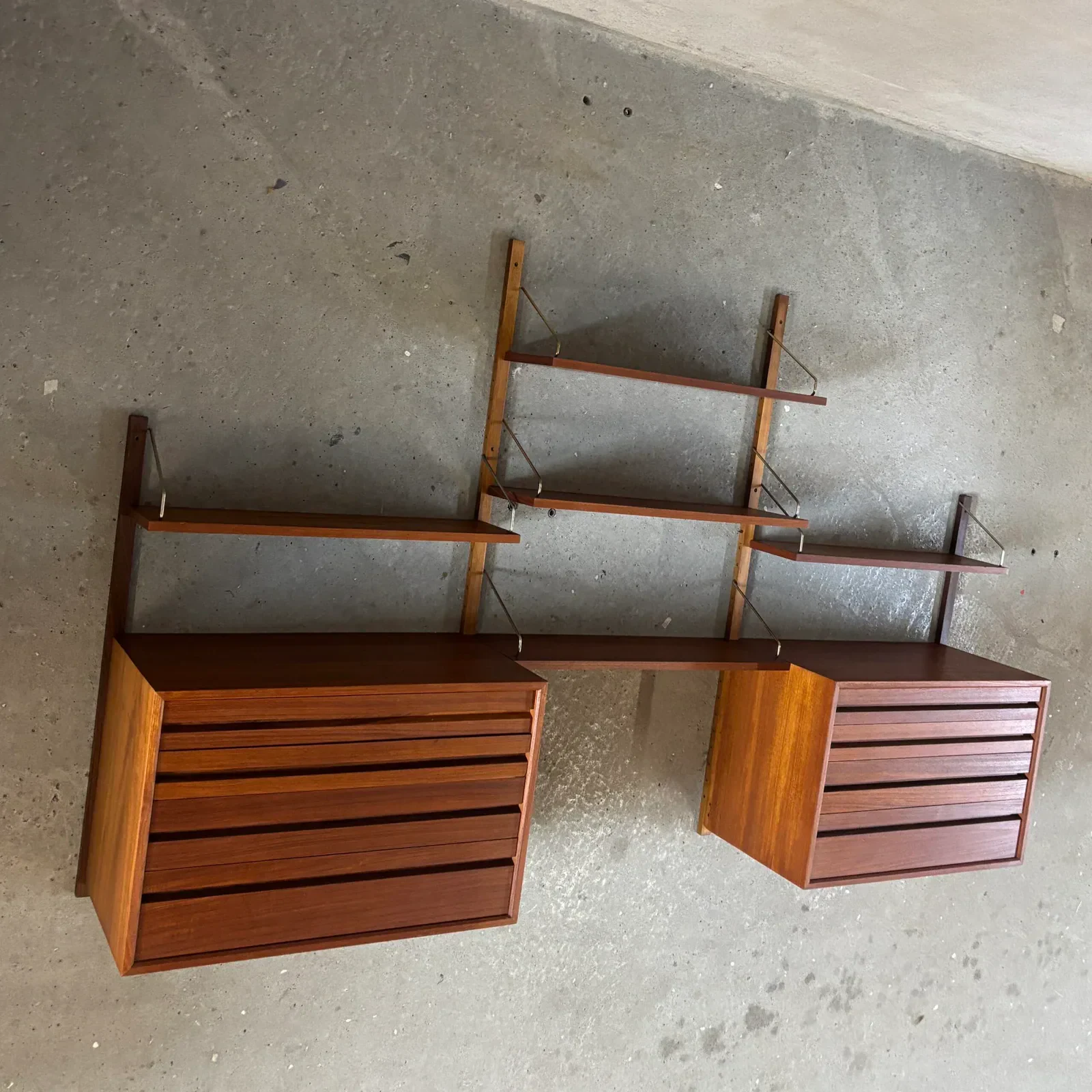 A three-bay wall system with vertical timber rails, thin metal hangers, open shelves and two suspended drawer cabinets against a concrete wall.