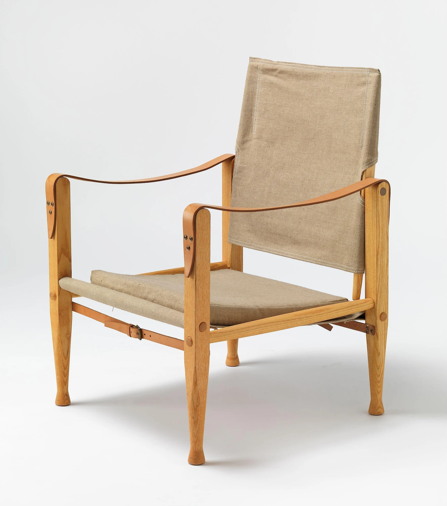 Three-quarter view of a pale timber Safari Chair with separate canvas seat and back, curved leather arm straps and exposed round-ended joints.