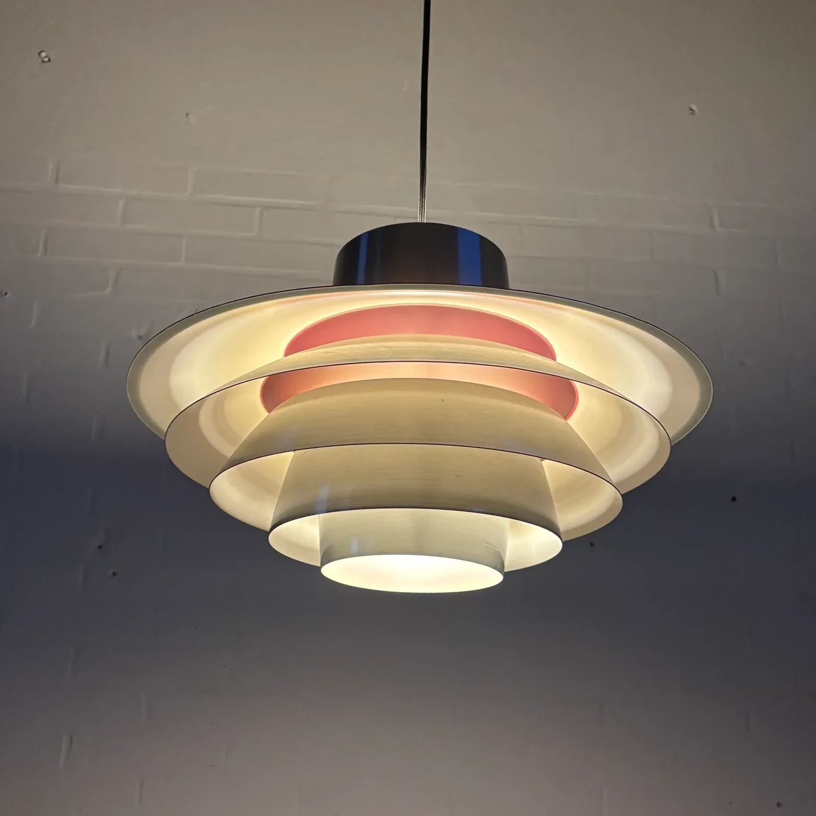 A lit pendant seen from slightly below has nested circular metal shades glowing around a concealed bulb beneath a dark cylindrical top cap.