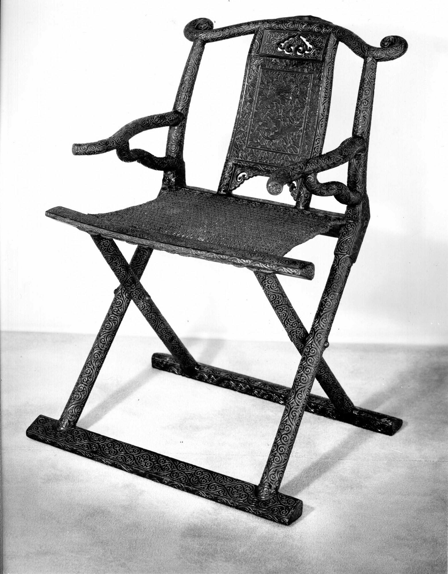 Full view of a Chinese carved-lacquer folding armchair with a rattan seat, crossed legs, long feet, curved arms and a bird-and-flower back panel.