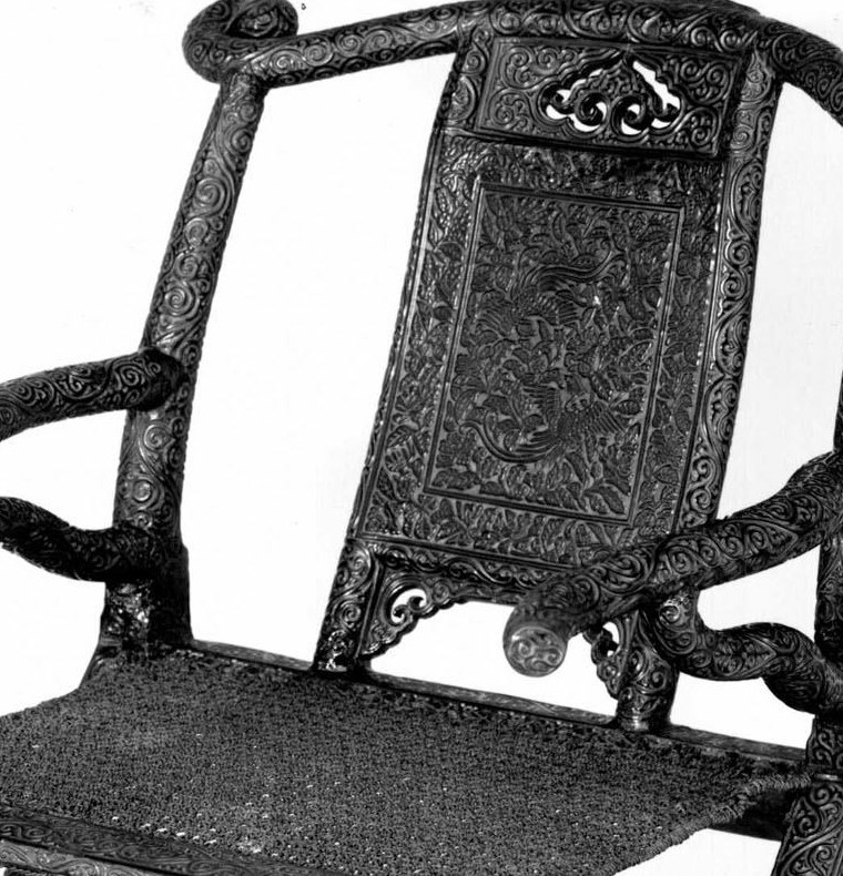 Close detail of the chair back showing two long-tailed birds among dense flowers inside carved borders, immediately above the woven rattan seat.