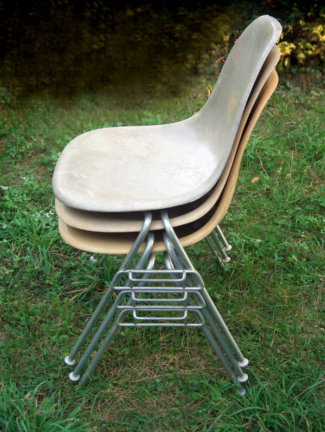 Three pale Eames shell side chairs stacked on wire bases, seen from the side on a lawn.