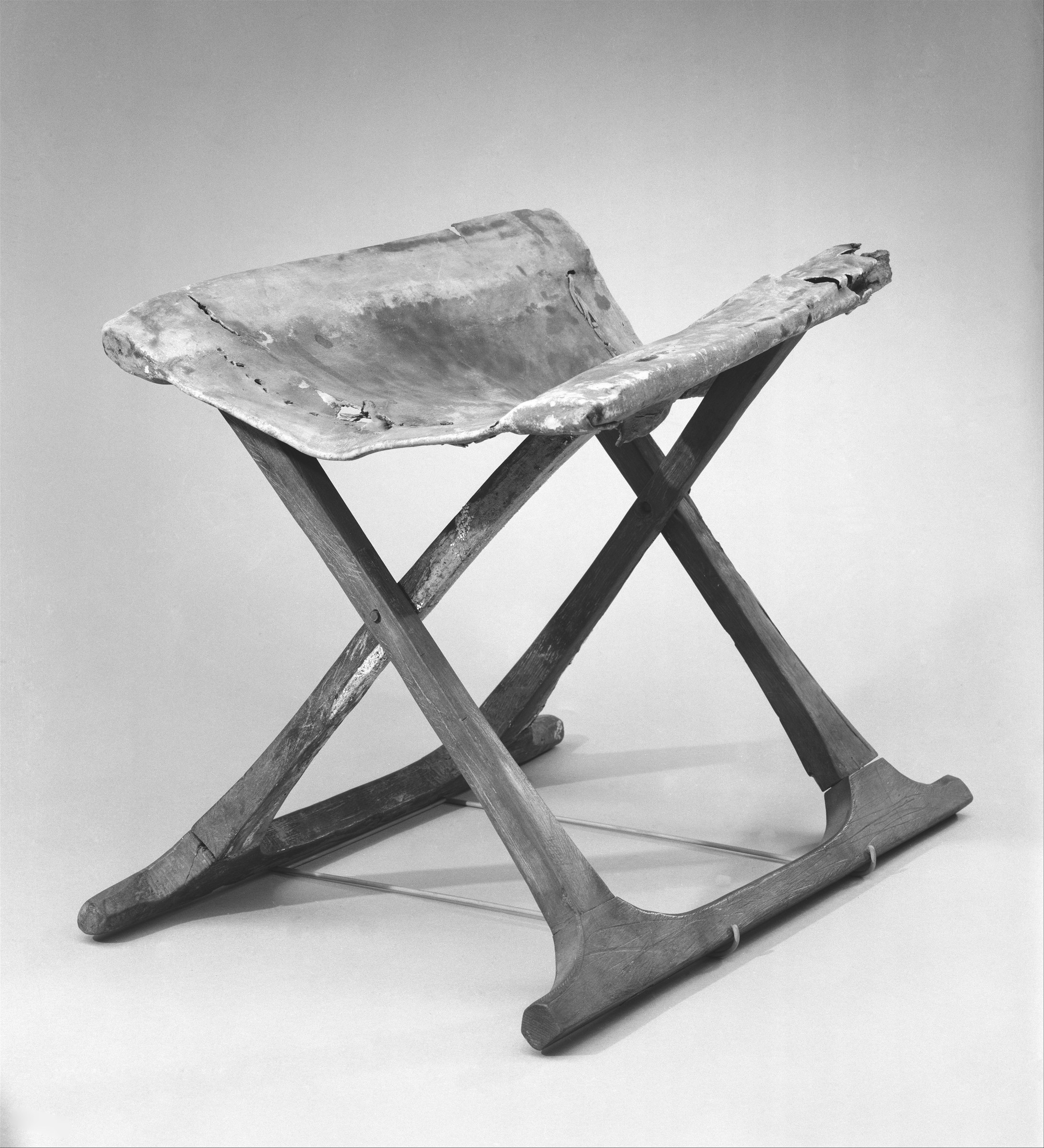 Three-quarter view of an ancient Egyptian folding stool with two crossed wooden end frames, long feet and a worn hide seat stretched between upper rails.