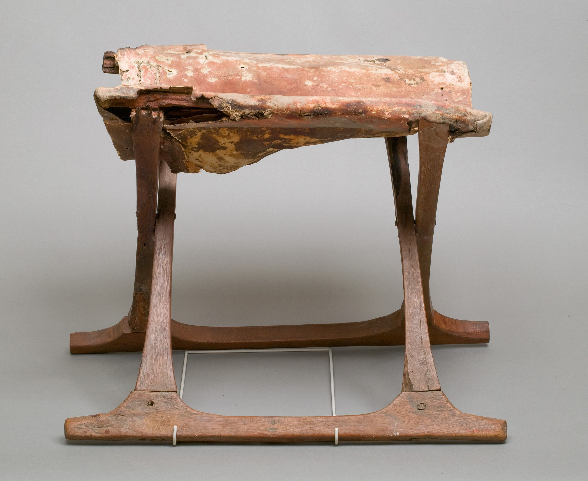 Long side view of the Egyptian folding stool in which the crossed members align visually beneath the worn reddish hide and paired longitudinal rails.