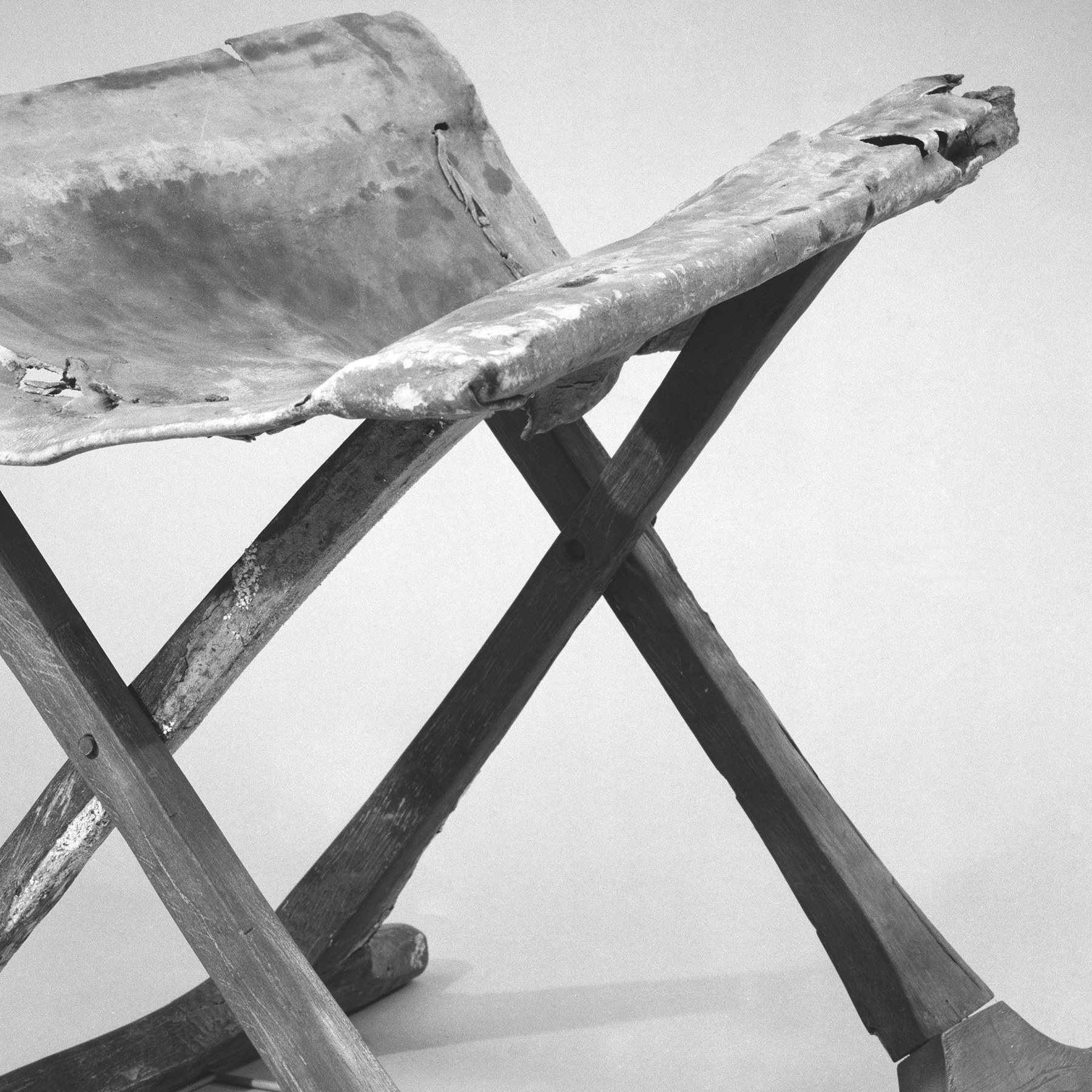 Close detail of the Egyptian stool showing cracked leather wrapped over an upper rail above crossing wooden members and their dark circular pivot points.