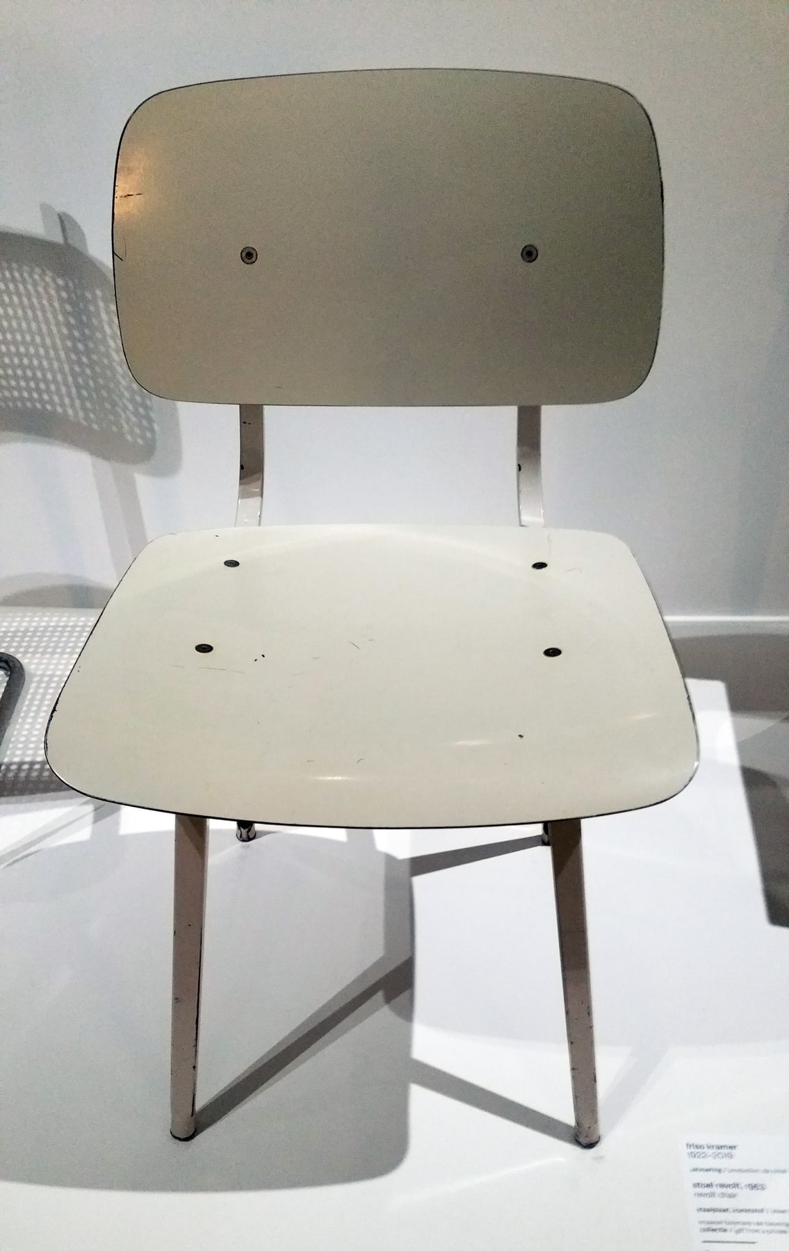 A Revolt chair with a pale separate seat and back on an open frame of folded sheet steel in a museum.