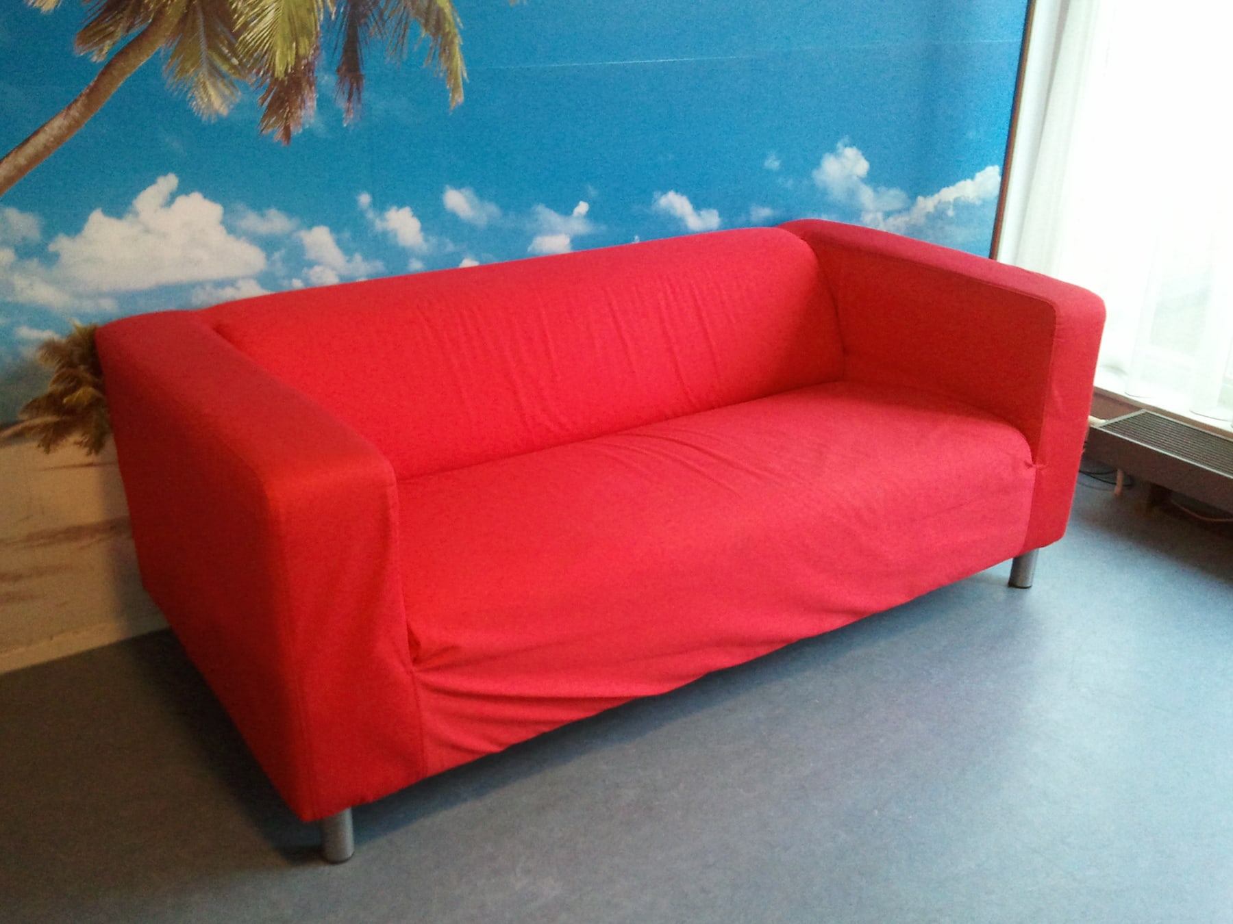 A compact red KLIPPAN sofa with low square arms, a continuous seat and a loose cover reaching below the base.