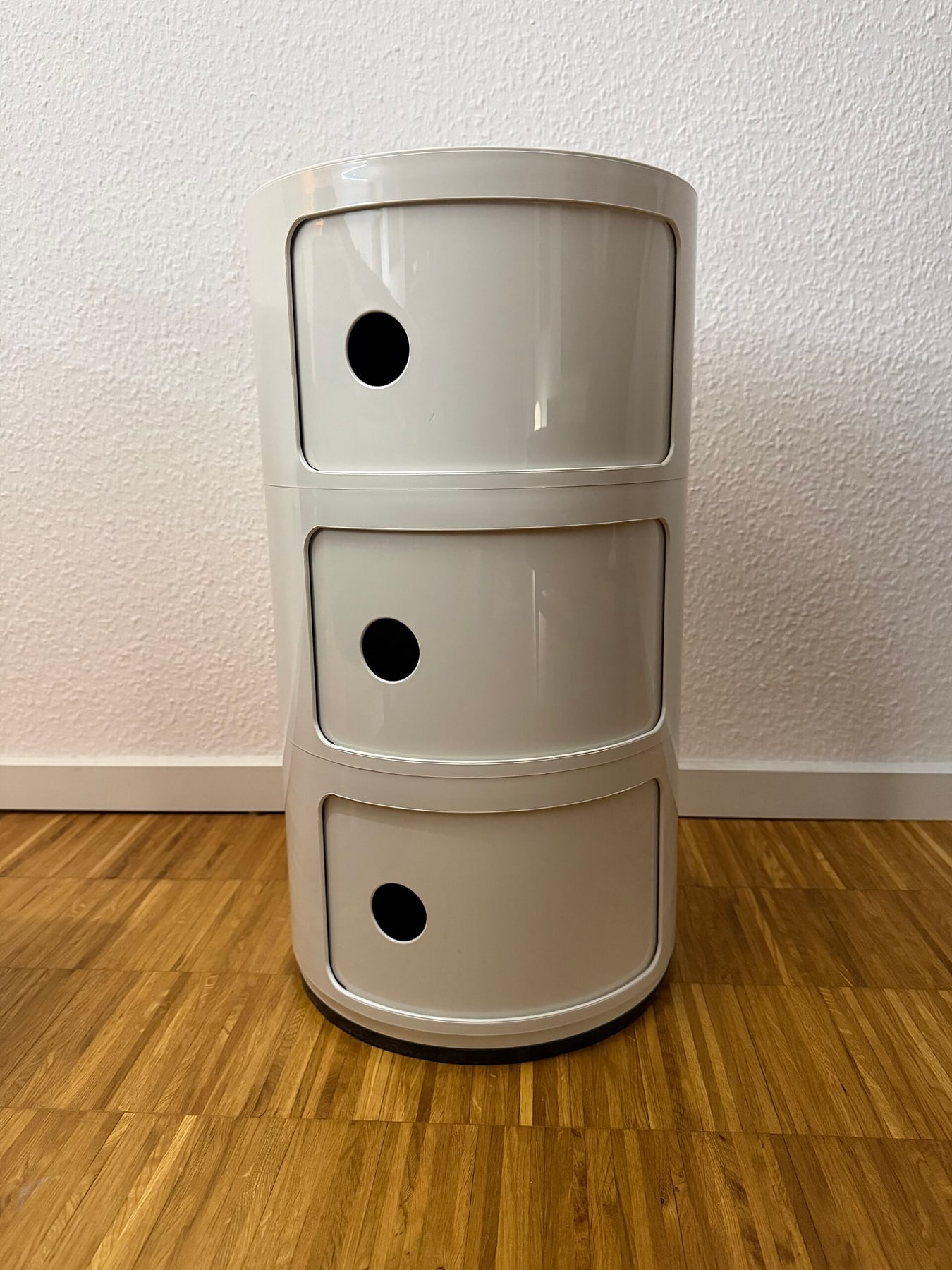 A white cylindrical Componibili cabinet with three sliding doors and round finger holes, standing on a parquet floor.