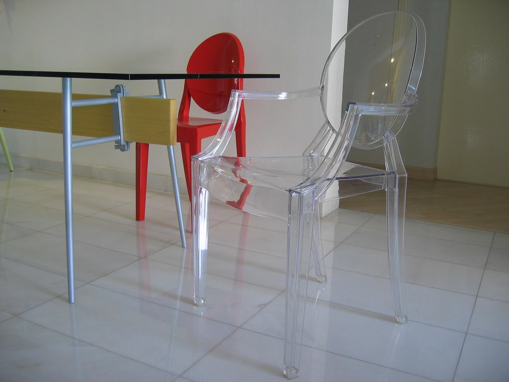 A clear Louis Ghost armchair beside a glass-topped table, with a red armless Victoria Ghost chair behind it.