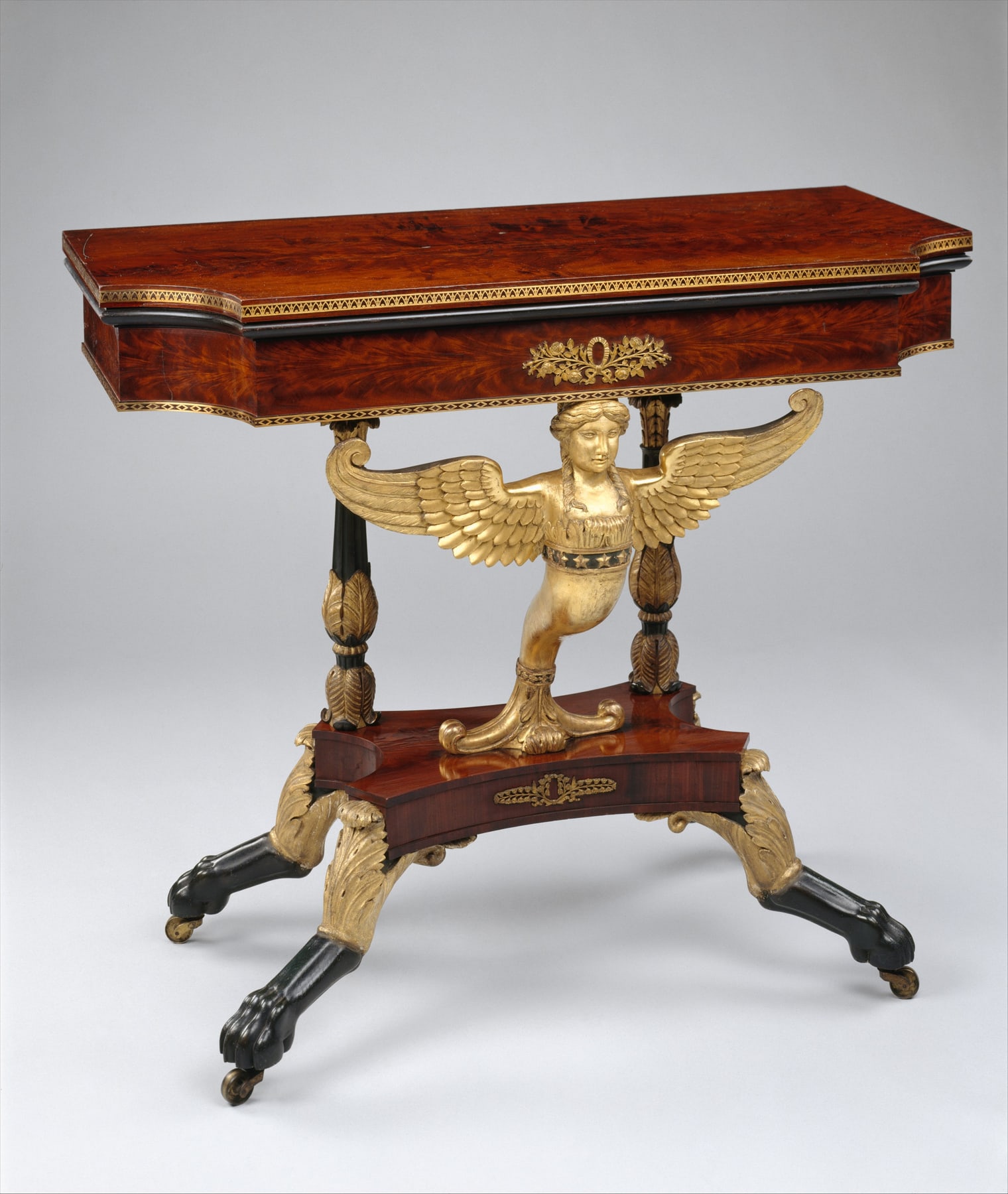 Lannuier’s card table with a folded top above a gilded winged figure and sculptural base.