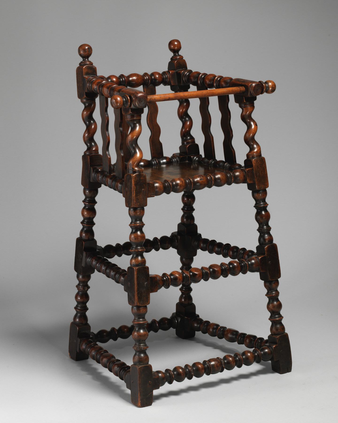 A walnut high chair has long turned legs beneath a compact seat surrounded by low wooden rails.