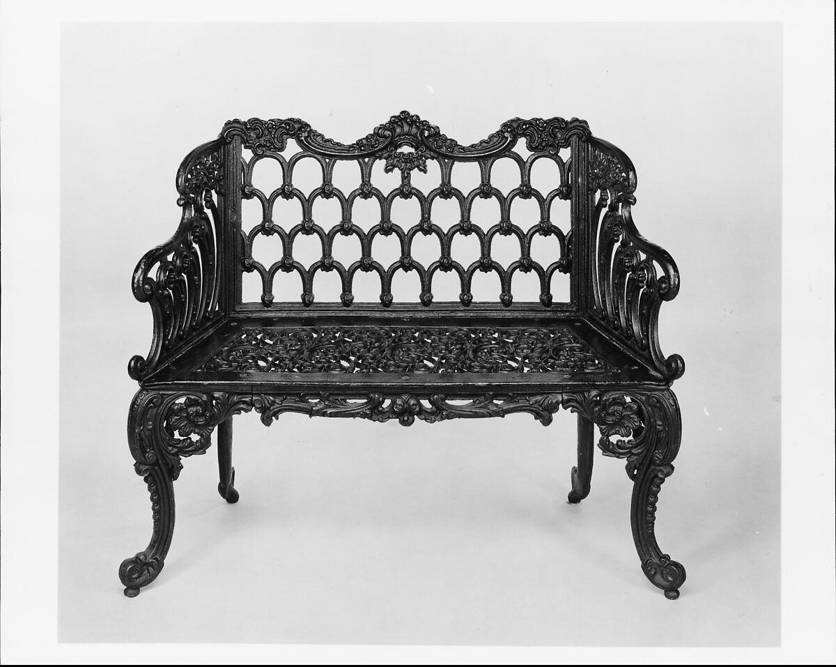 A dark cast-iron bench has an openwork back of repeated curved shapes, a perforated seat and densely ornamented arms and legs.