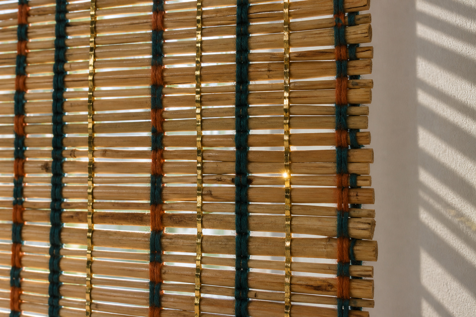 A woven blind combines uneven horizontal reeds with teal, rust and metallic-looking threads, admitting patches of warm daylight onto the wall behind.