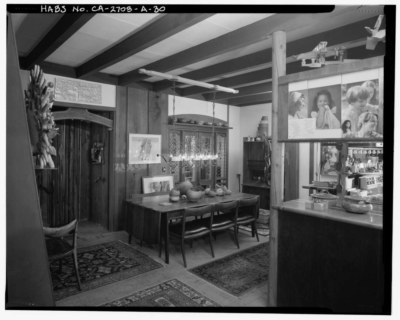 A black-and-white survey view of the Maloof dining area, where wooden chairs and a table sit among the possessions of a furnished home.