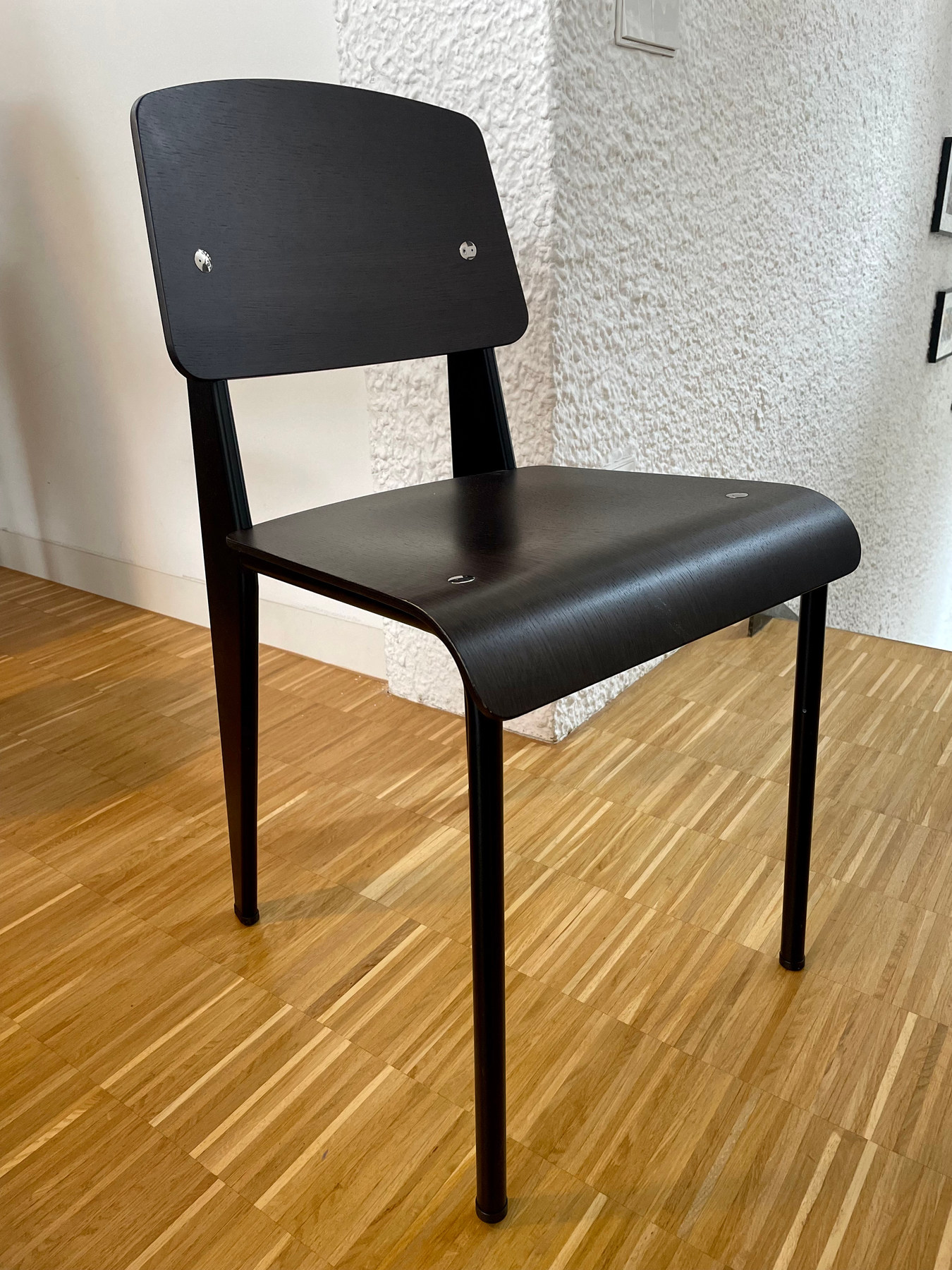 Front three-quarter view of a black wooden chair with curved seat and back, exposed fasteners, slender round front legs and deeper steel rear supports.
