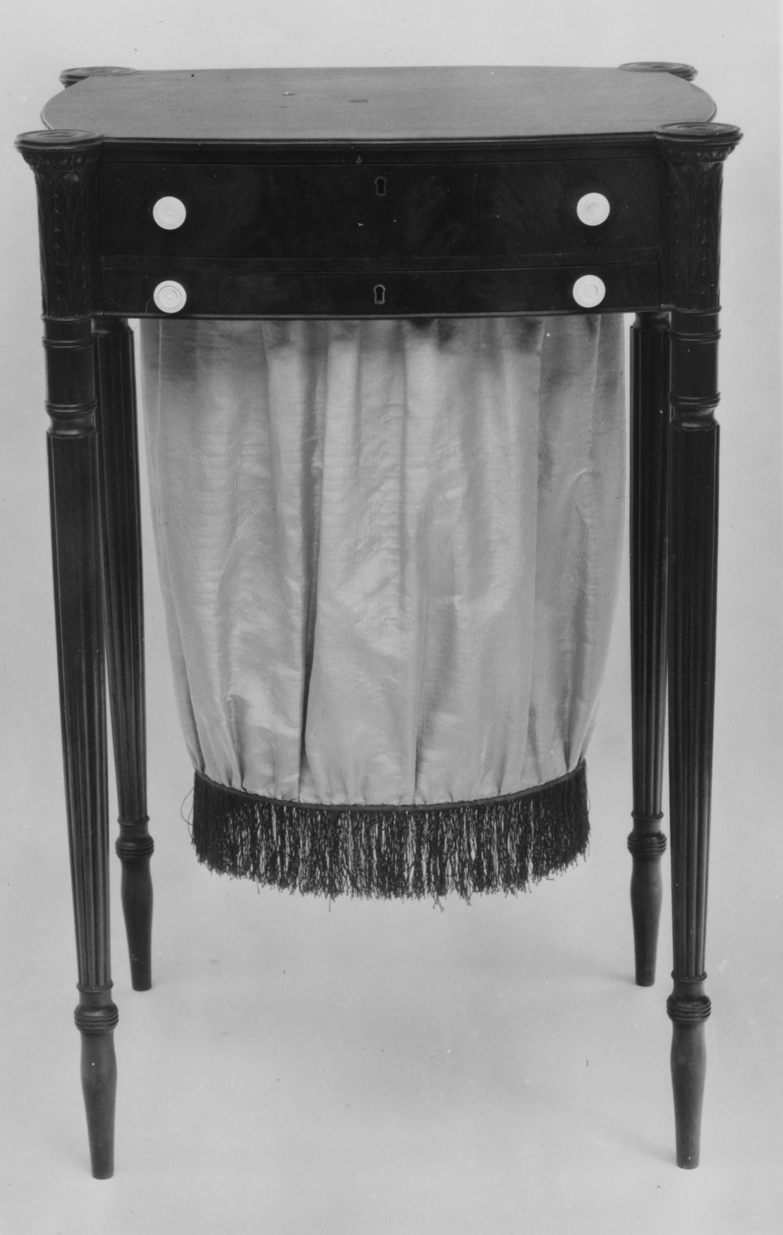 A small wooden worktable on slender legs has drawers above a deep gathered fabric bag.
