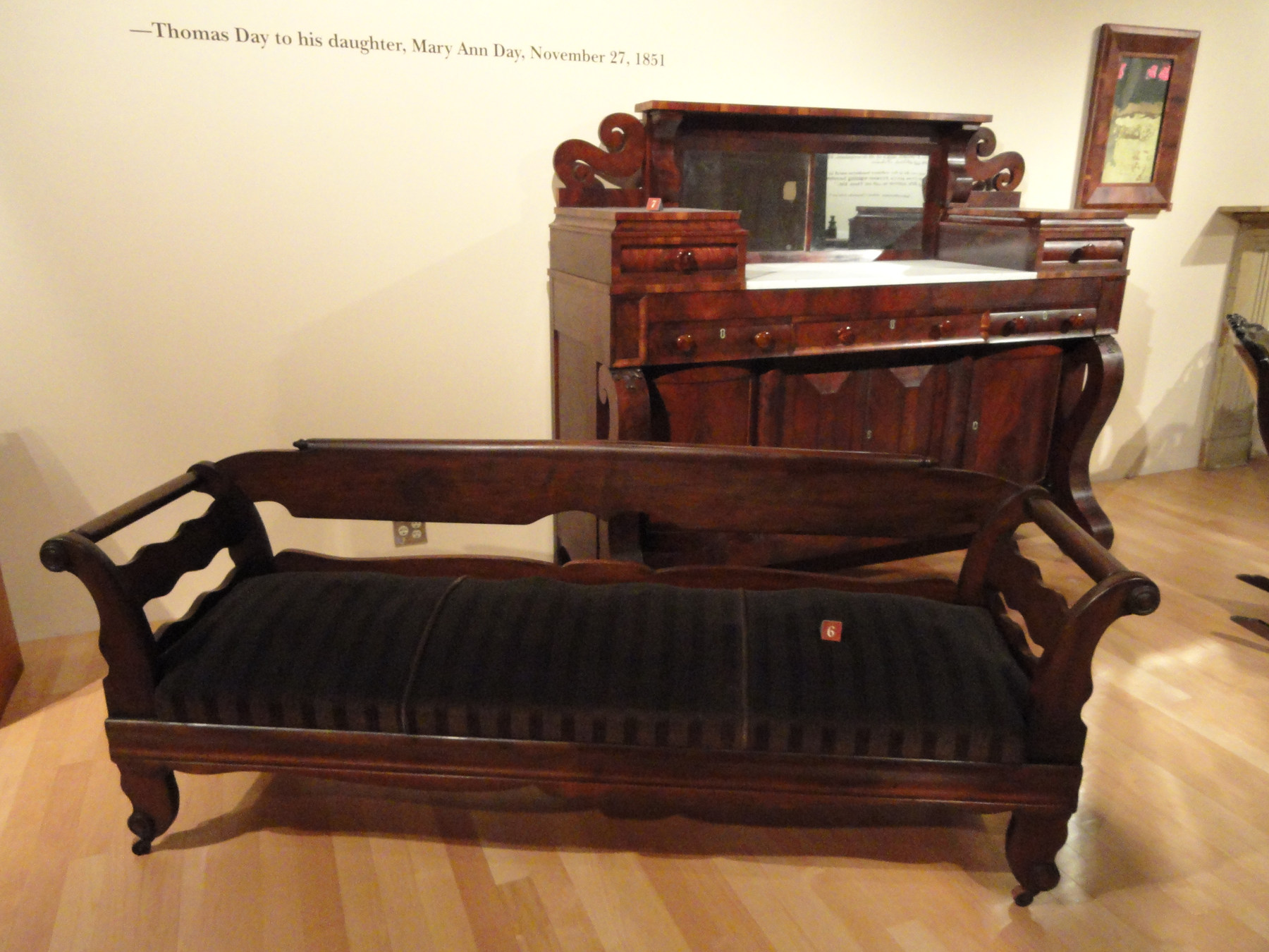 A dark wooden settee with curved arms stands in front of storage furniture with prominent scrolls in a museum display.