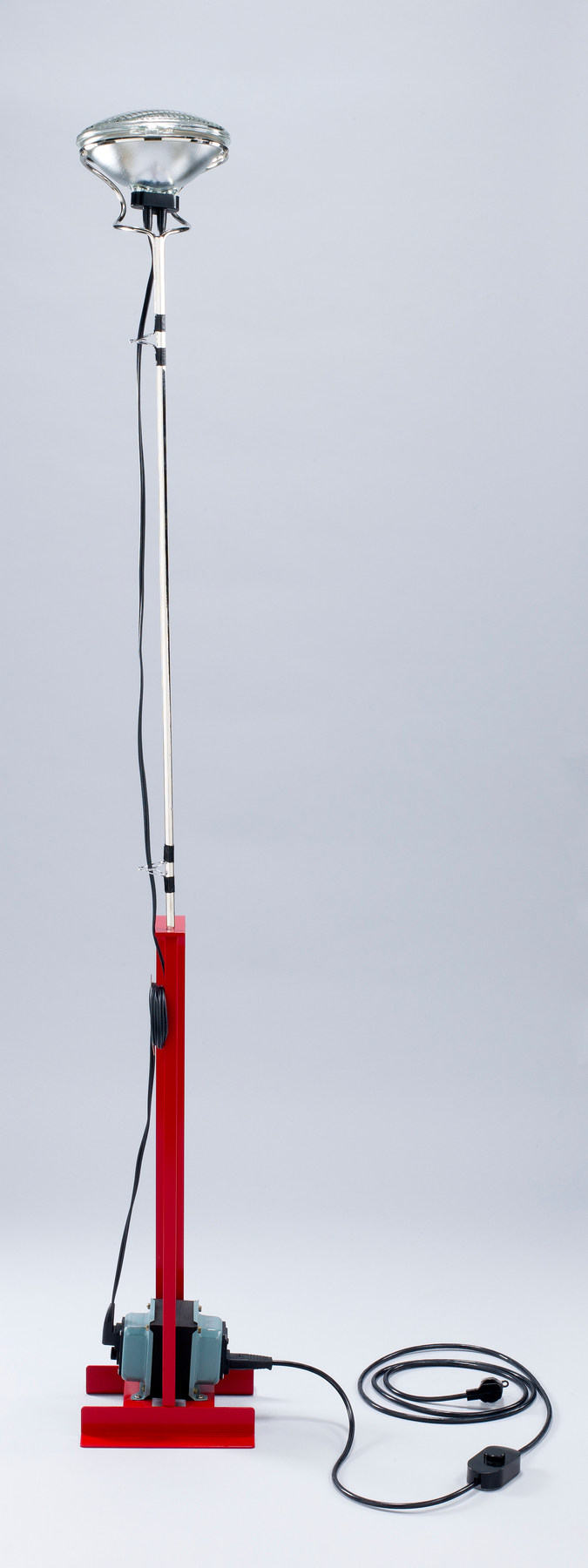 A slender upright lamp has an upward-facing headlight, visible cable guides and a red base carrying its transformer.
