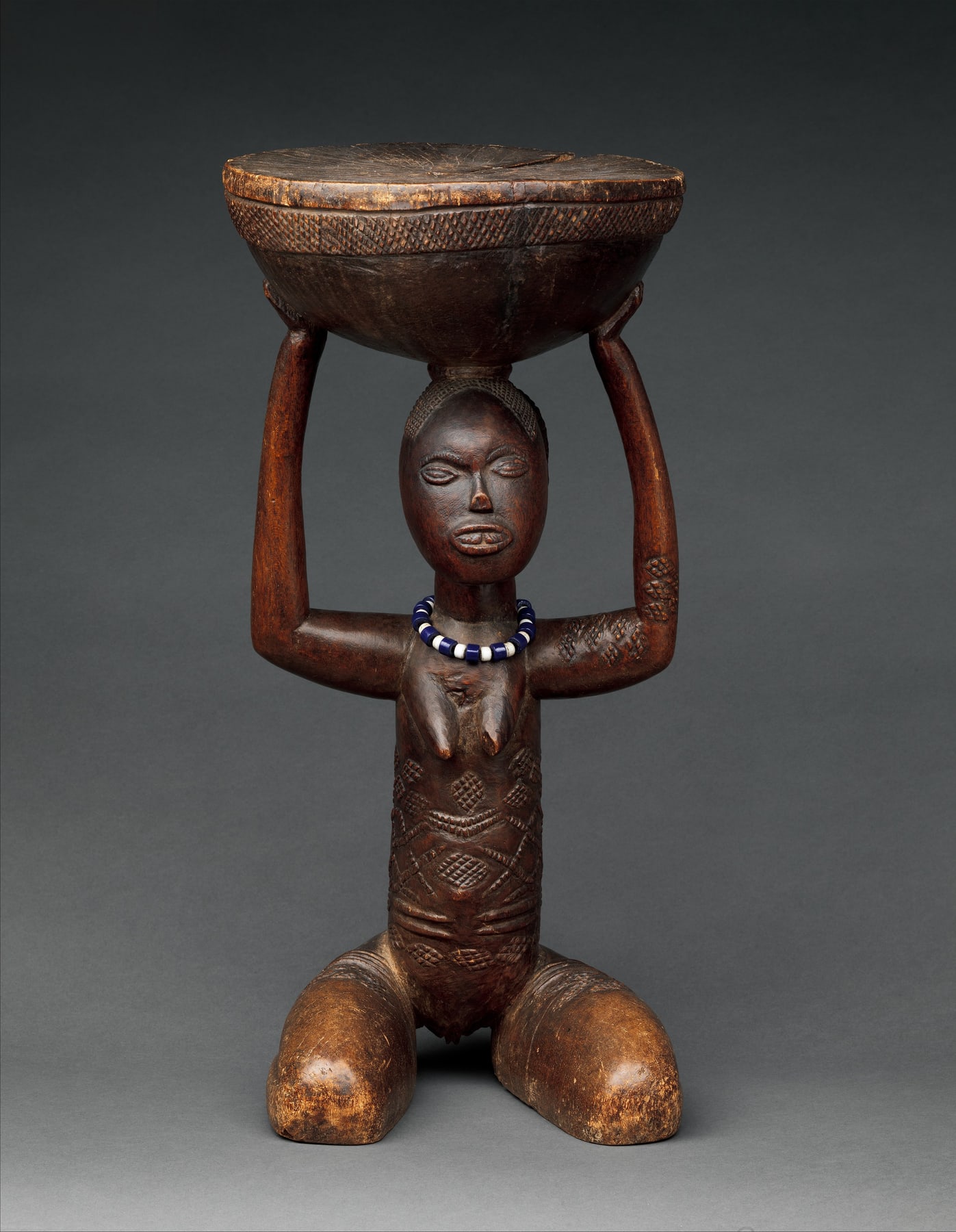 Luba royal seat with a kneeling female figure supporting a circular top and a band of blue and white beads.