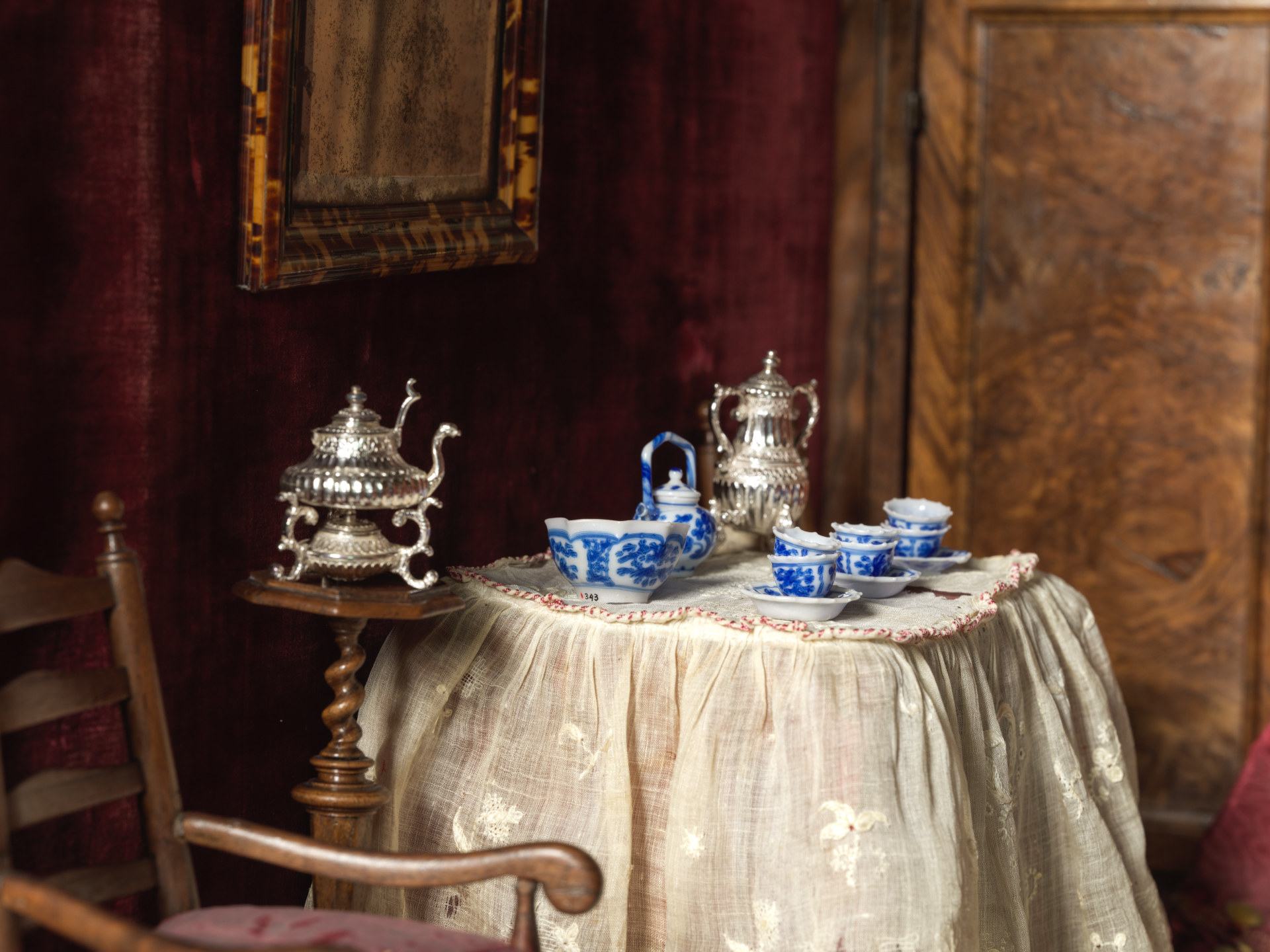 Miniature table in the lying-in room set with blue-and-white vessels, silver pots and embroidered linen beside a wooden chair.