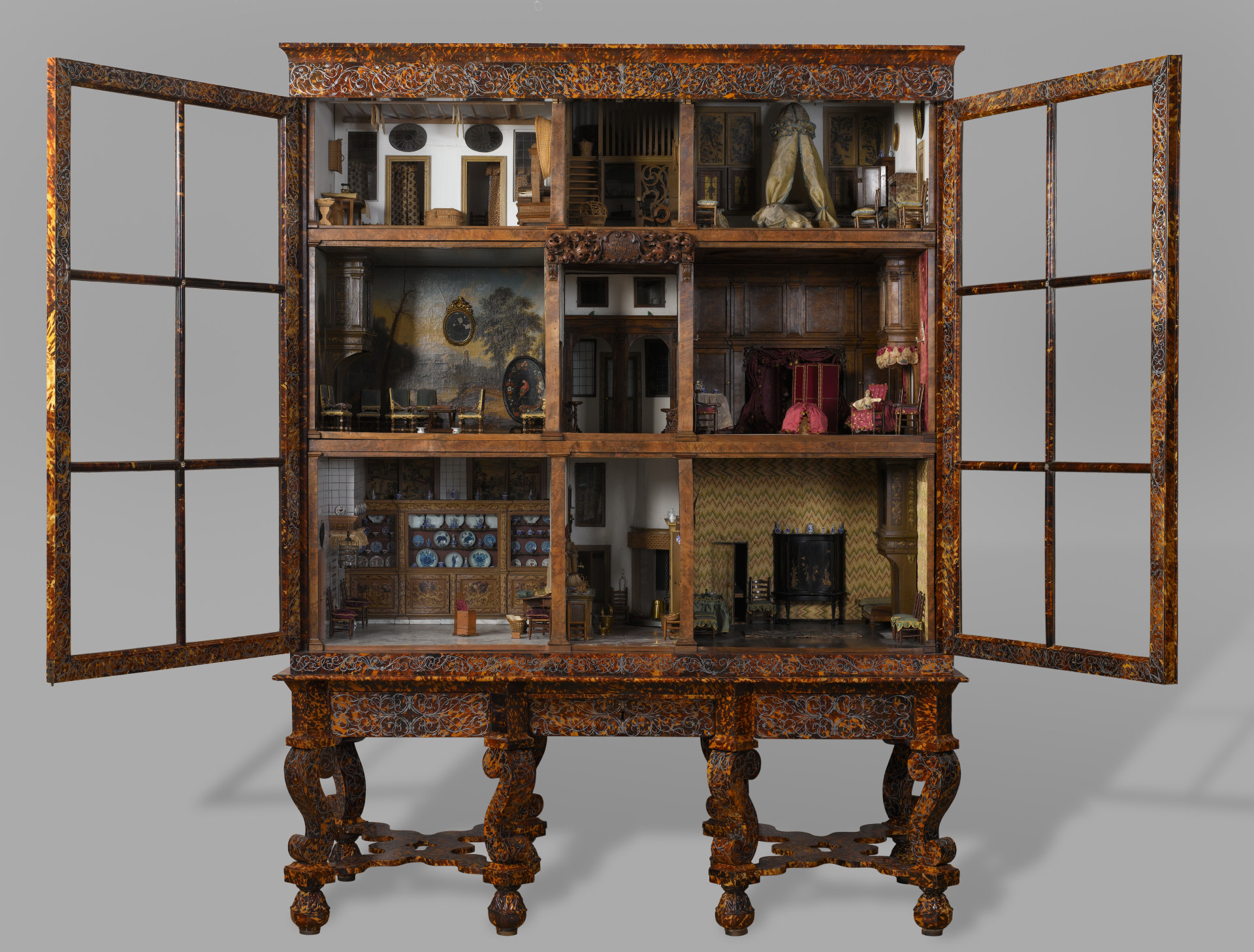 Open tortoiseshell-veneered cabinet with two glazed doors framing three tiers of furnished miniature rooms on a carved stand.