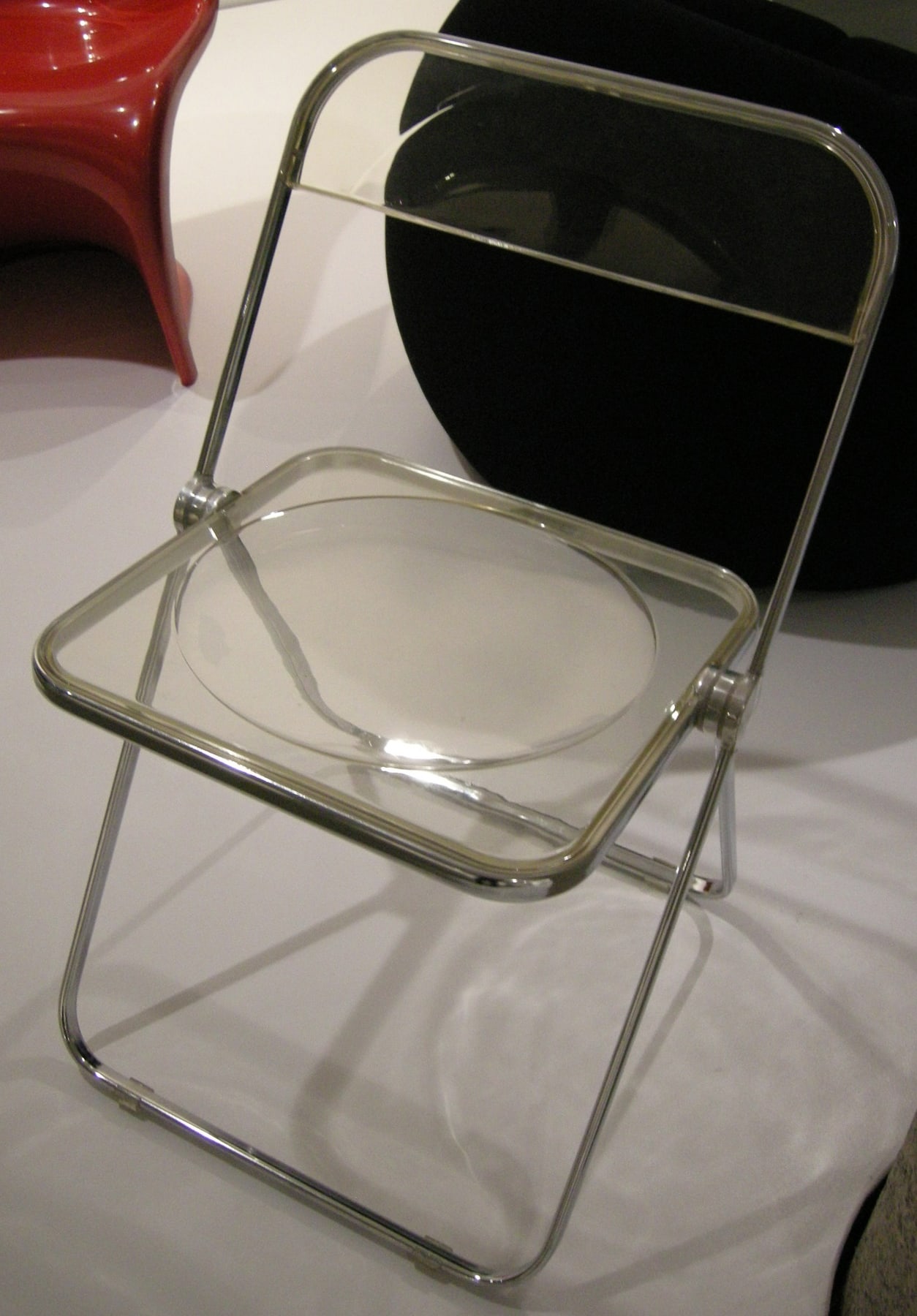 A transparent Plia folding chair with a tubular metal frame, clear seat and clear rectangular back in a museum display.