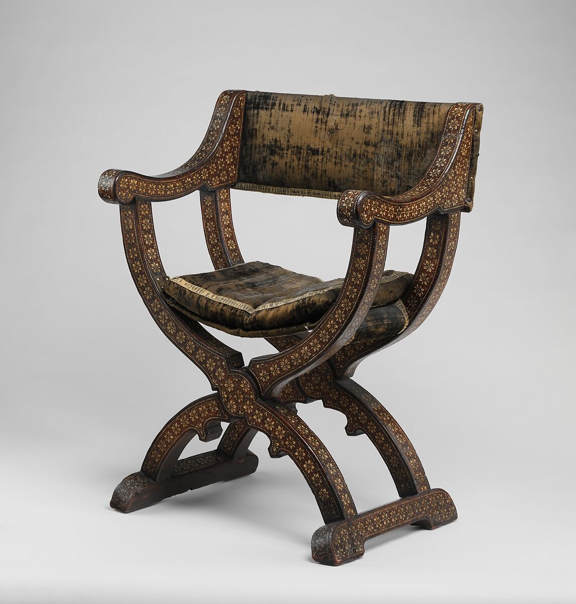 Three-quarter studio view of a richly inlaid Spanish hip-joint armchair with crossed curved supports, runner-like feet and worn dark textile.