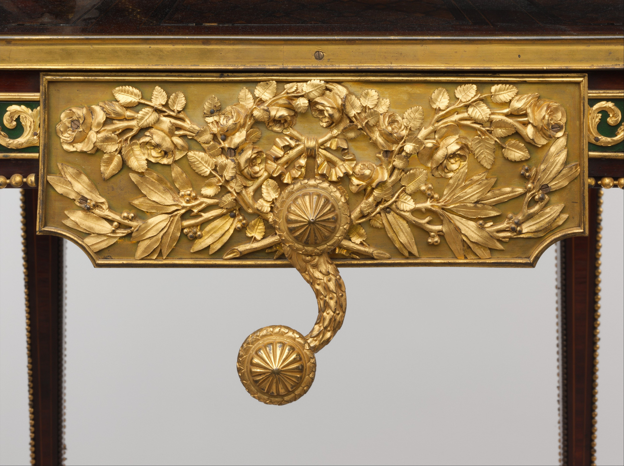 Close view of a gilded floral plaque beneath the table edge with an S-shaped detachable crank hanging from its centre.