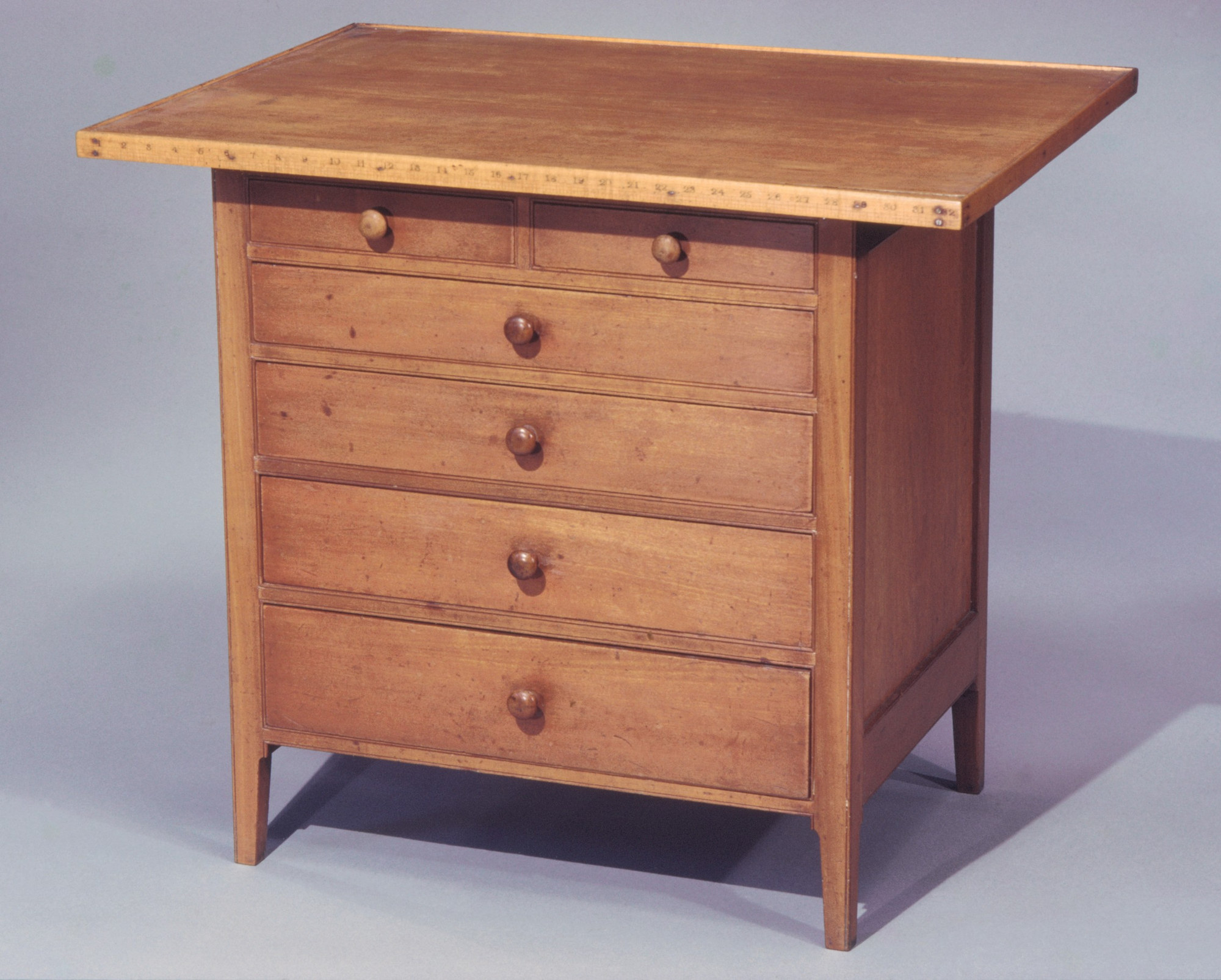 Slightly lower view of the same Shaker sewing table, showing its numbered worktop edge, plain side panel, wooden knobs and tapered legs.
