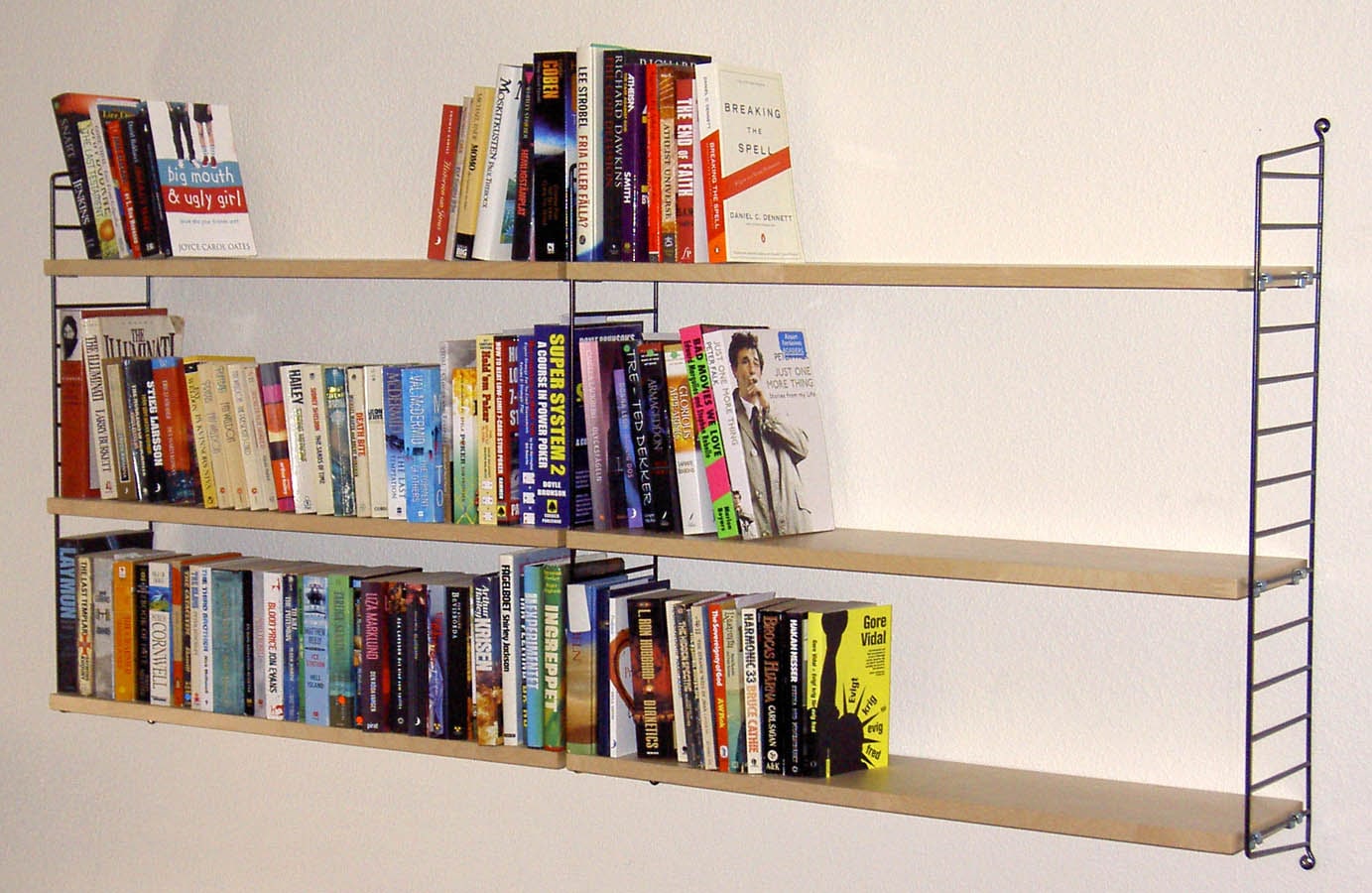 Two bays of String shelving with thin black wire sides and three pale wooden shelves, partly filled with books on a white wall.