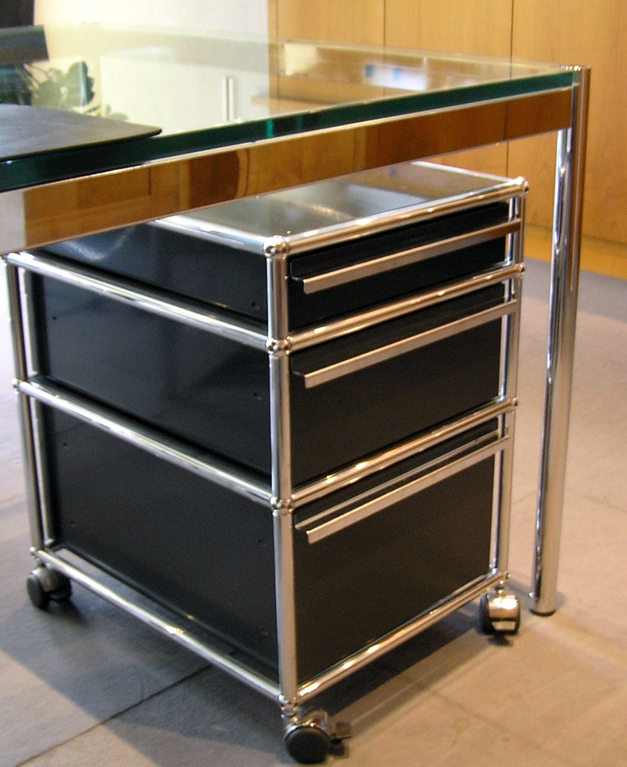 A dark USM Haller drawer unit on castors under a glass desk, with shiny tubular edges and round metal connectors.