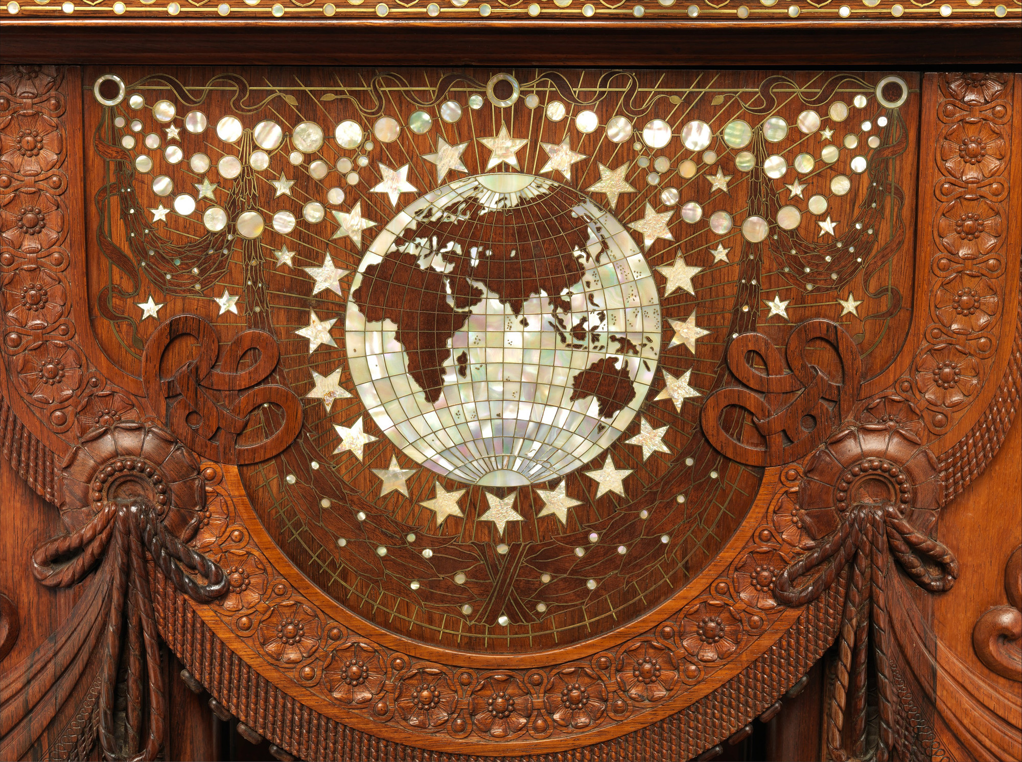 Close view of a shell-inlaid globe surrounded by stars, brass lines, carved flowers and hanging drapery at the table end.
