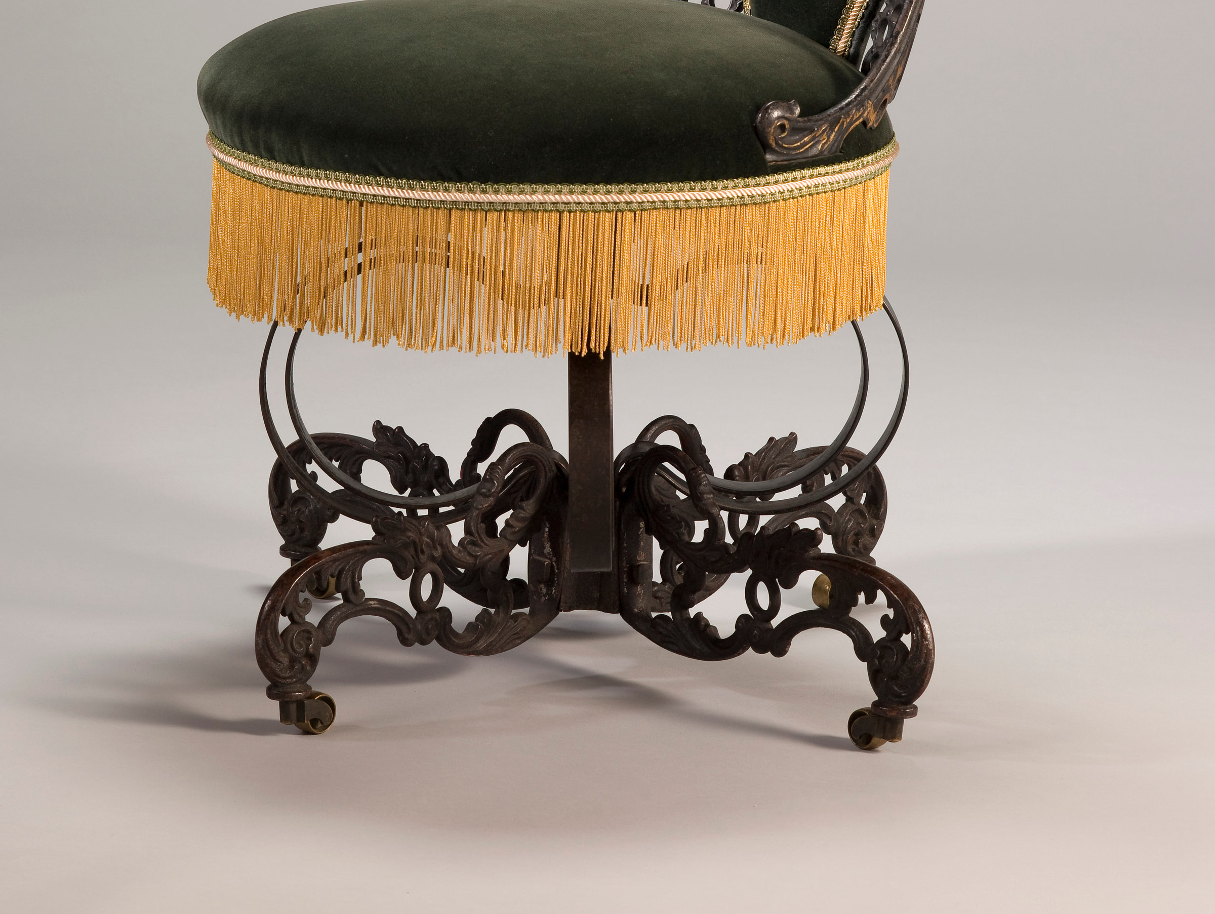 Detail of Warren's chair base showing gold fringe above a central metal post, large bowed steel elements, cast scrollwork and small brass casters.