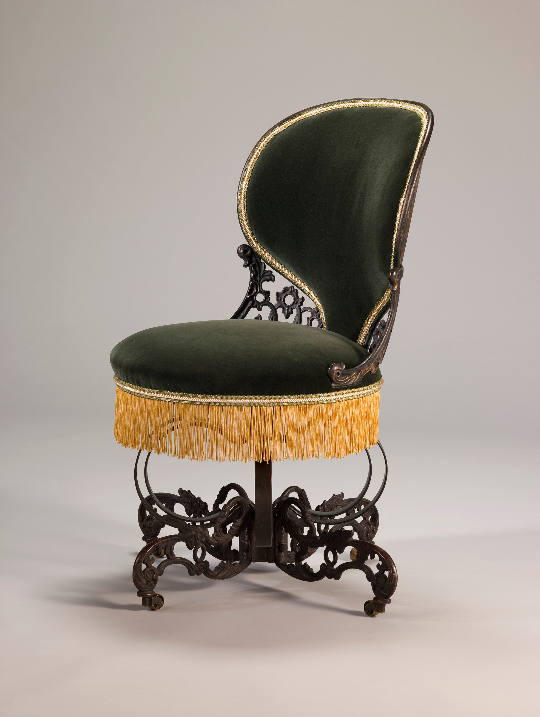 Three-quarter view of Warren's Centripetal Spring Chair with dark green velvet, gold fringe, ornamental iron scrollwork, a central support and casters.
