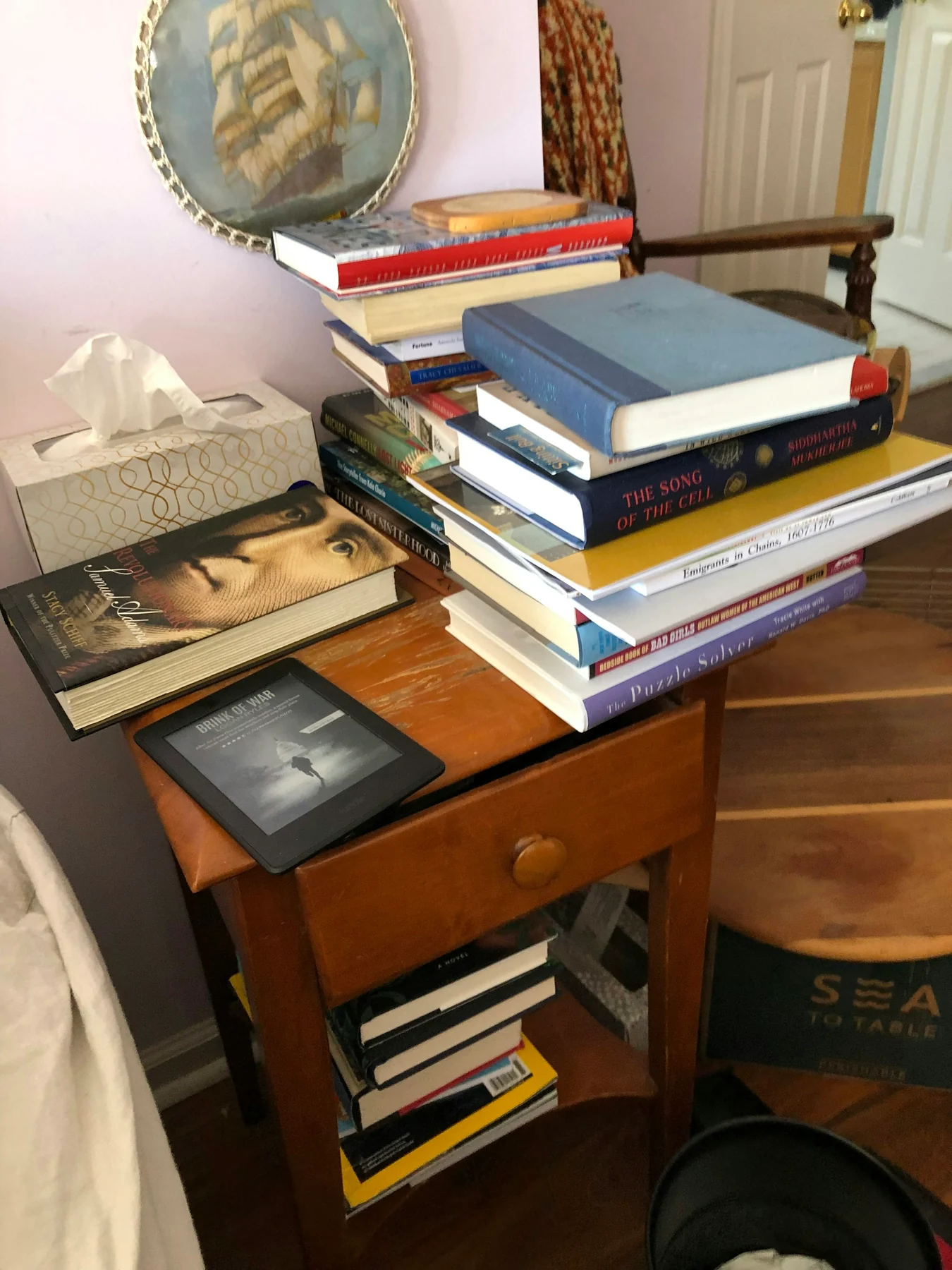Wooden bedside table crowded with books, an e-reader and a lamp, with more books on the lower shelf.