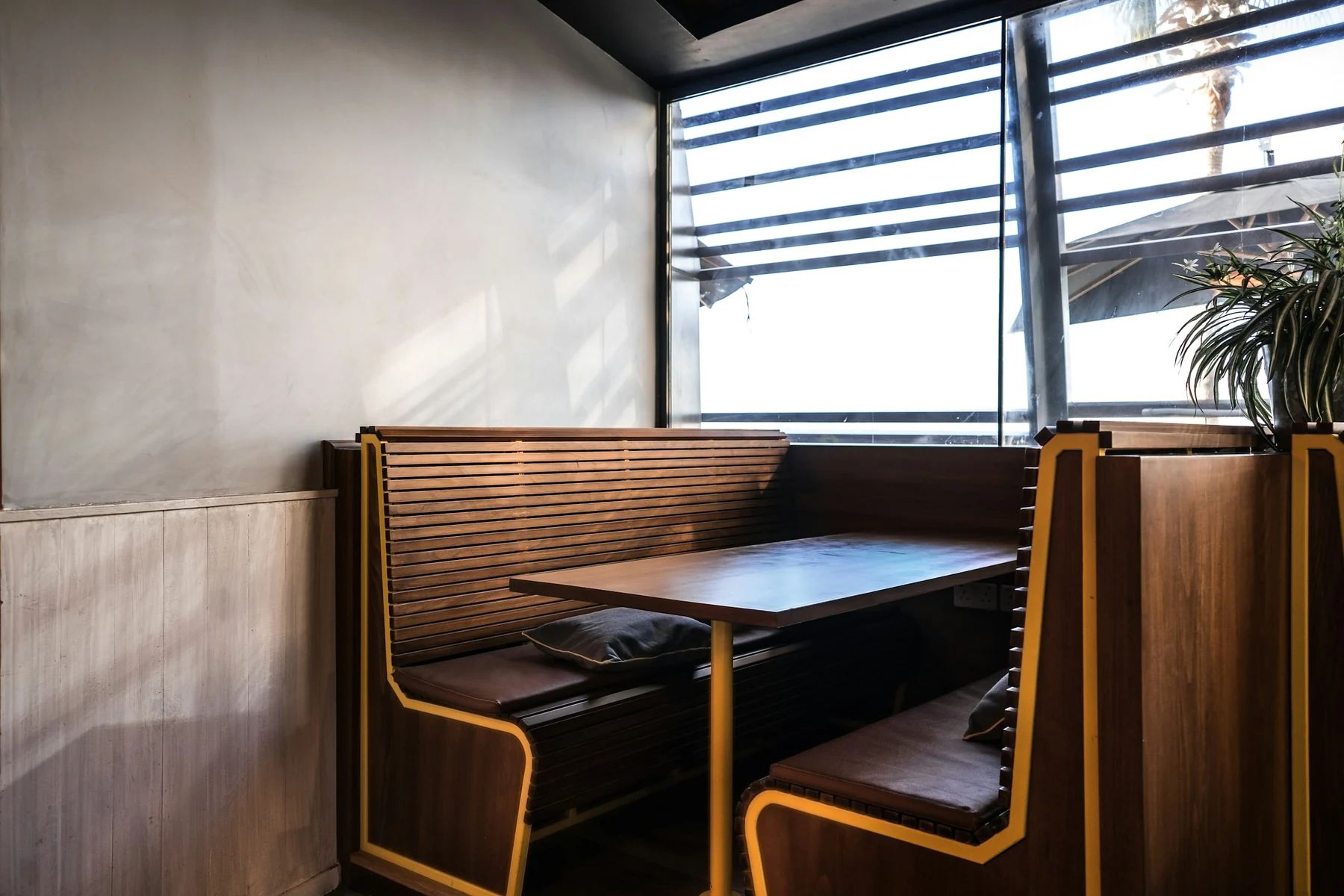 Restaurant table between two backed wooden benches with seat cushions, next to a wall and window.