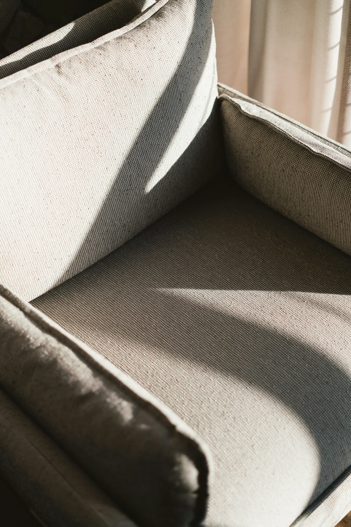 Close view of a pale upholstered armchair showing its curved inner back, seat, arms and piped edges in sunlight.