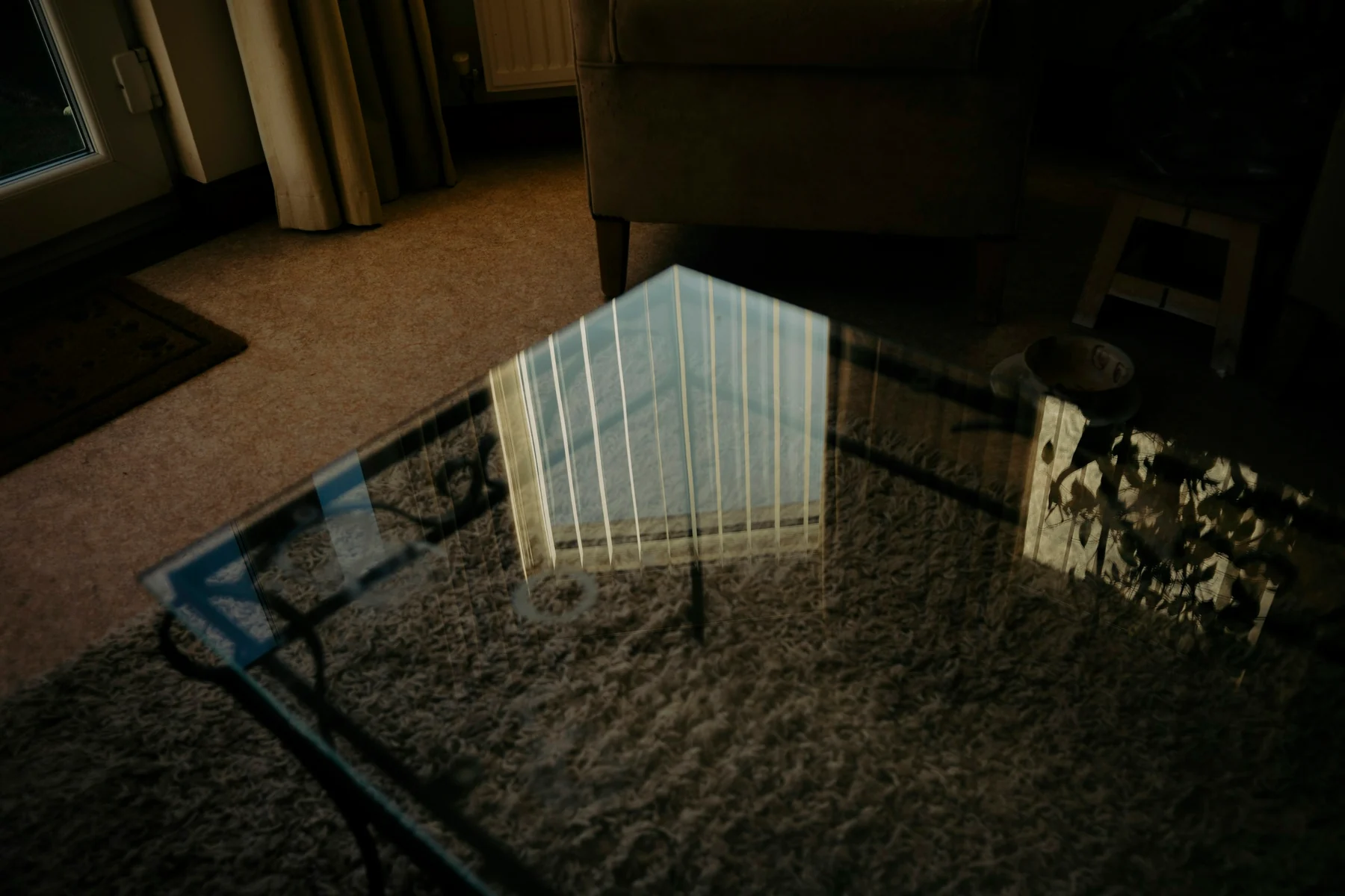 Glass coffee table reflecting a bright window and vertical lines above a rug and beside seating.