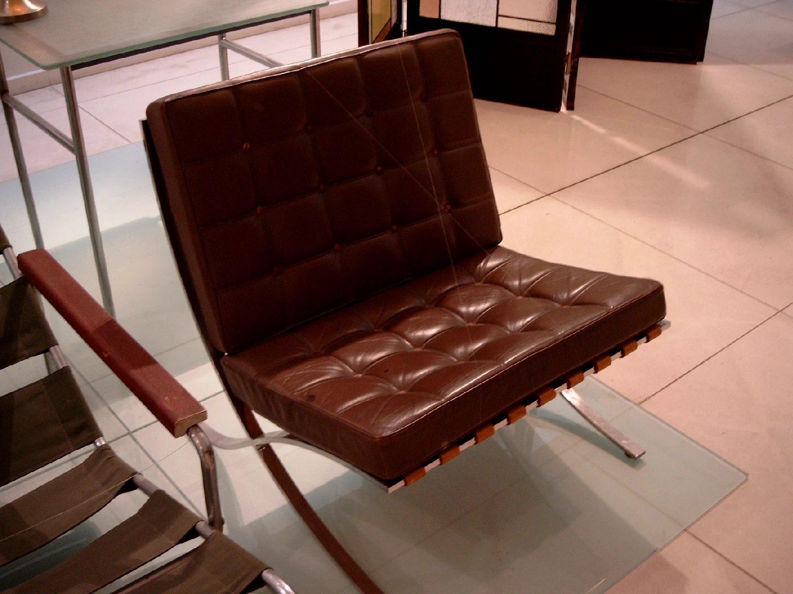 A brown leather Barcelona Chair photographed from the front and side, showing quilted cushions and part of the supporting strap arrangement.
