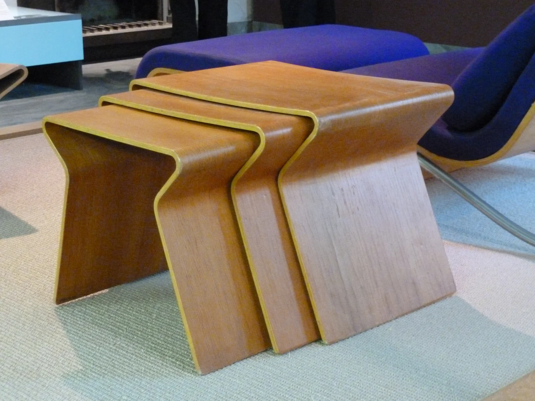 Three Grete Jalk nesting tables in graduated sizes, each made as a broad timber-coloured sheet that curves down at both ends, displayed with other furniture in a museum.