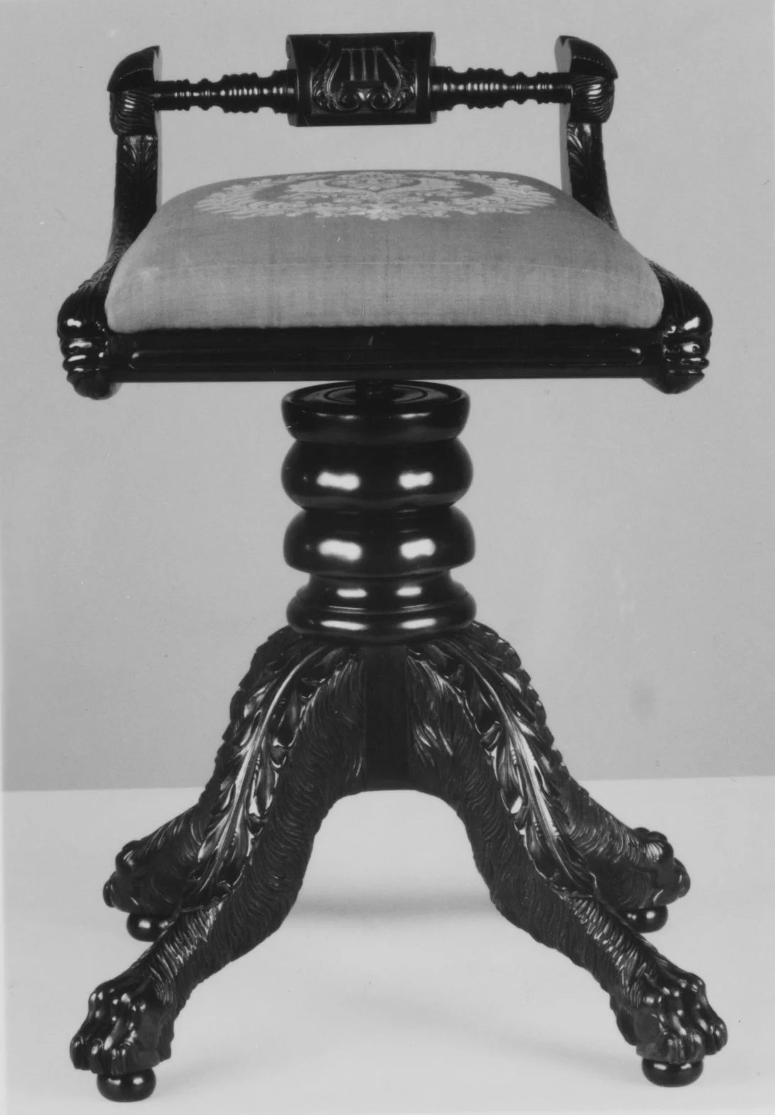 Black-and-white museum photograph of a piano stool with an upholstered seat, small back, central pedestal and four carved legs.